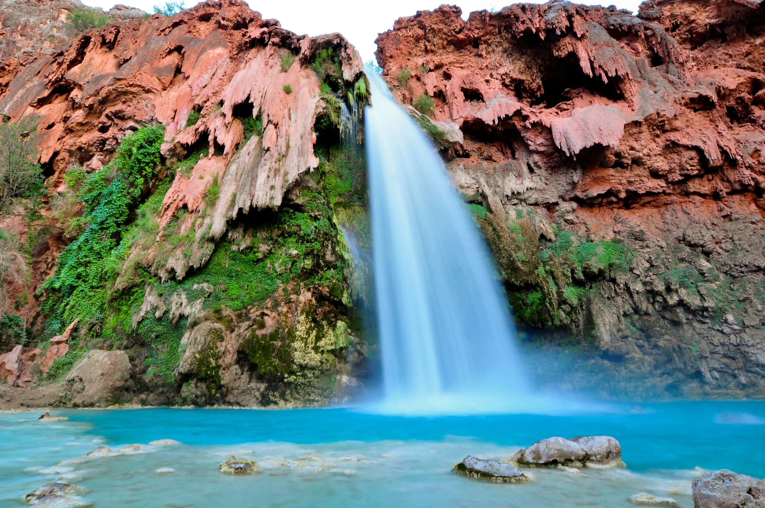 A waterfall cascading down red and green rocky cliffs into a turquoise pool of water surrounded by rocks