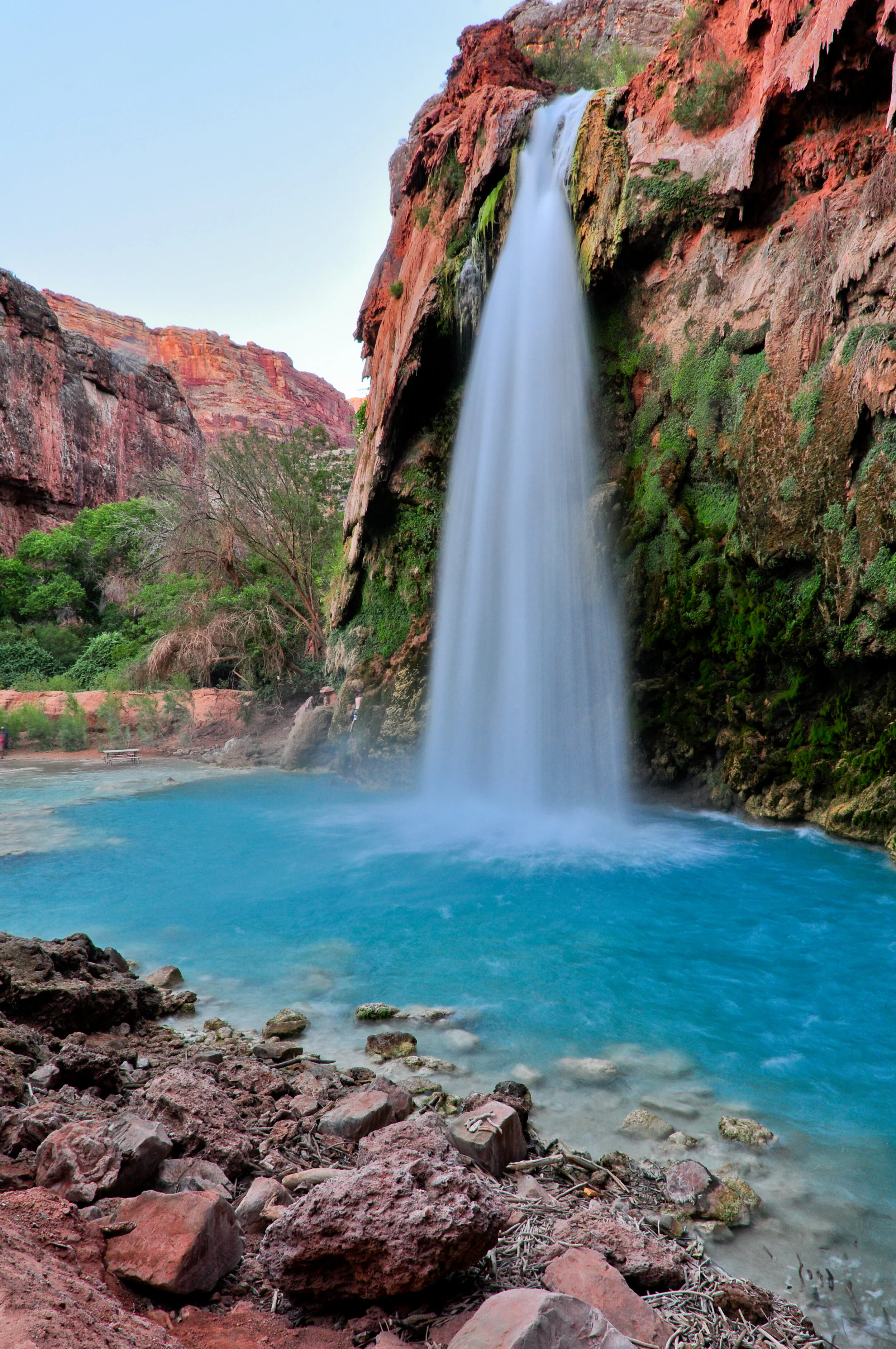 A tall waterfall cascading into a pool of blue water, surrounded by rocky terrain and green trees in a canyon with red and brown cliffs.