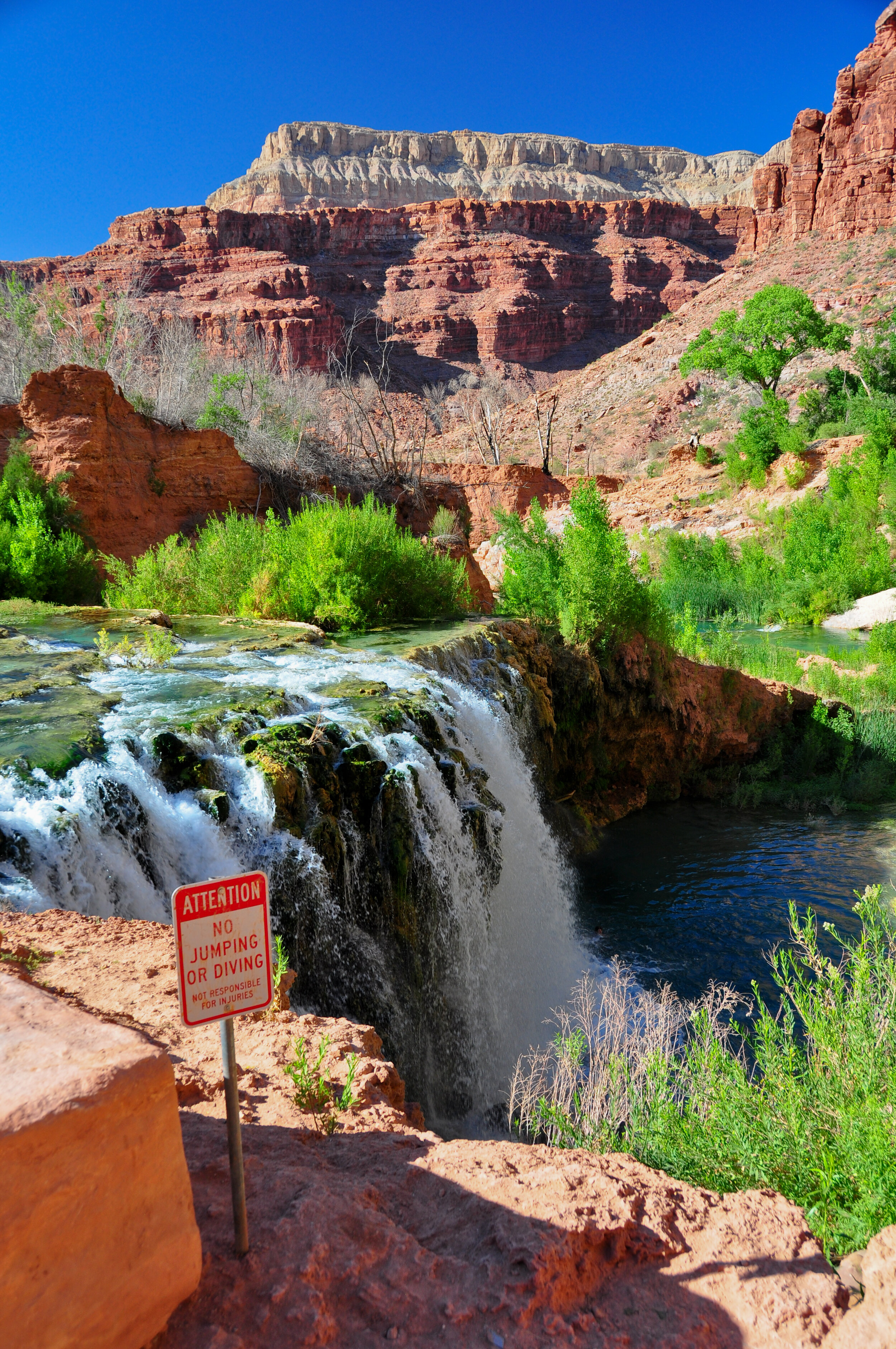 Waterfall with lush green trees and red rock formations in the background, and a sign that reads 'ATTENTION: NO JUMPING OR DIVING NOT RESPONSIBLE FOR INJURIES'.