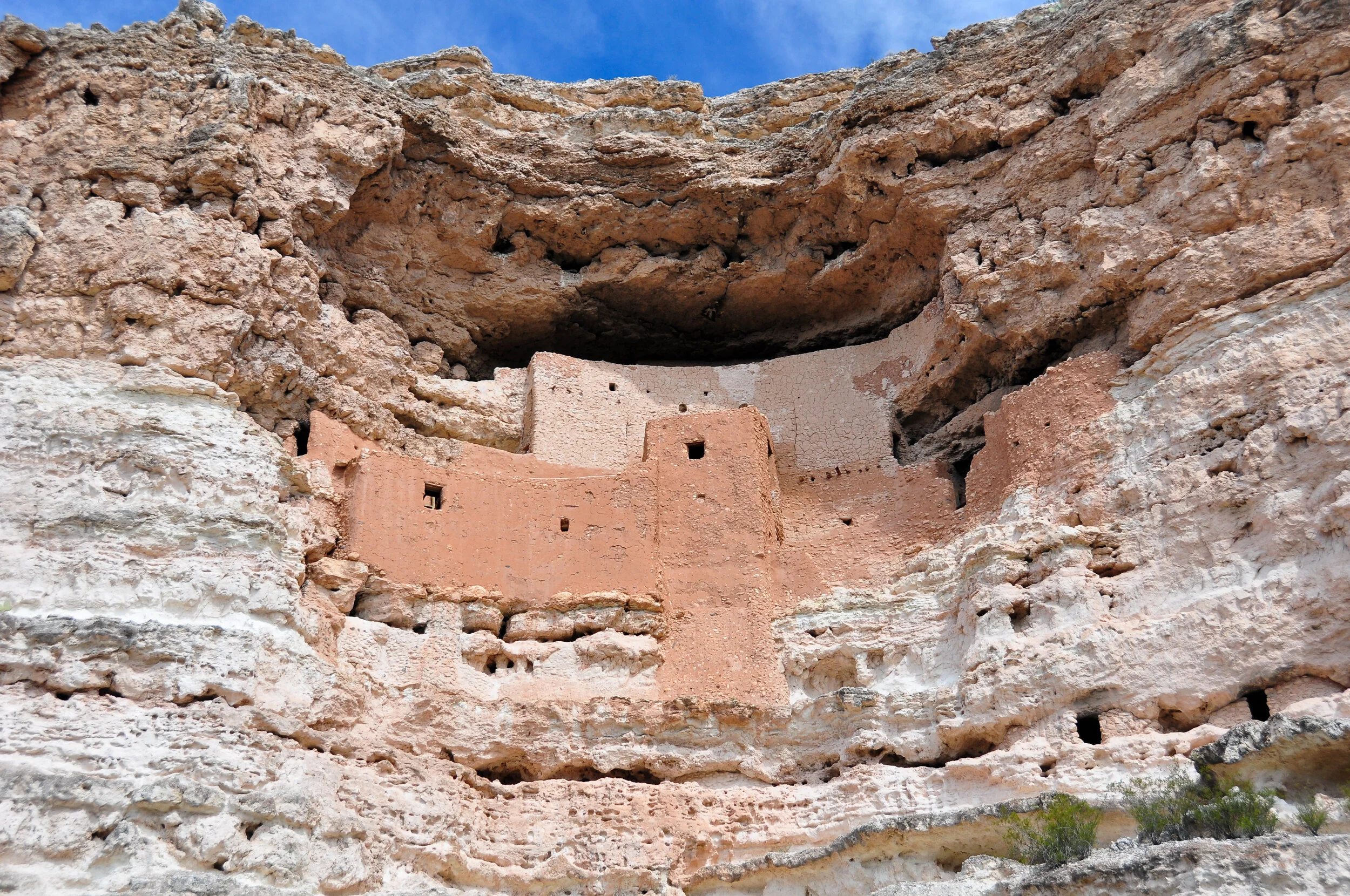 Ancient cliff dwellings built into a canyon wall with small windows, in a desert landscape with blue sky.