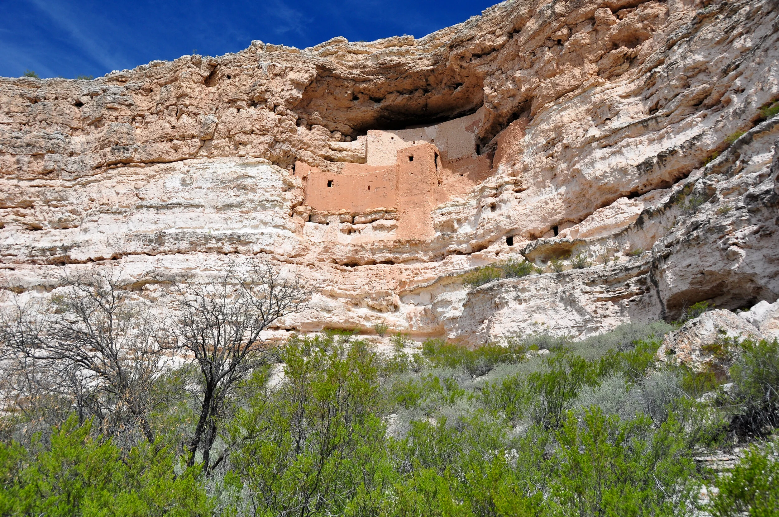 Ancient cliff dwelling built into the side of a rocky canyon wall, with sparse green vegetation at the base and a clear blue sky above.