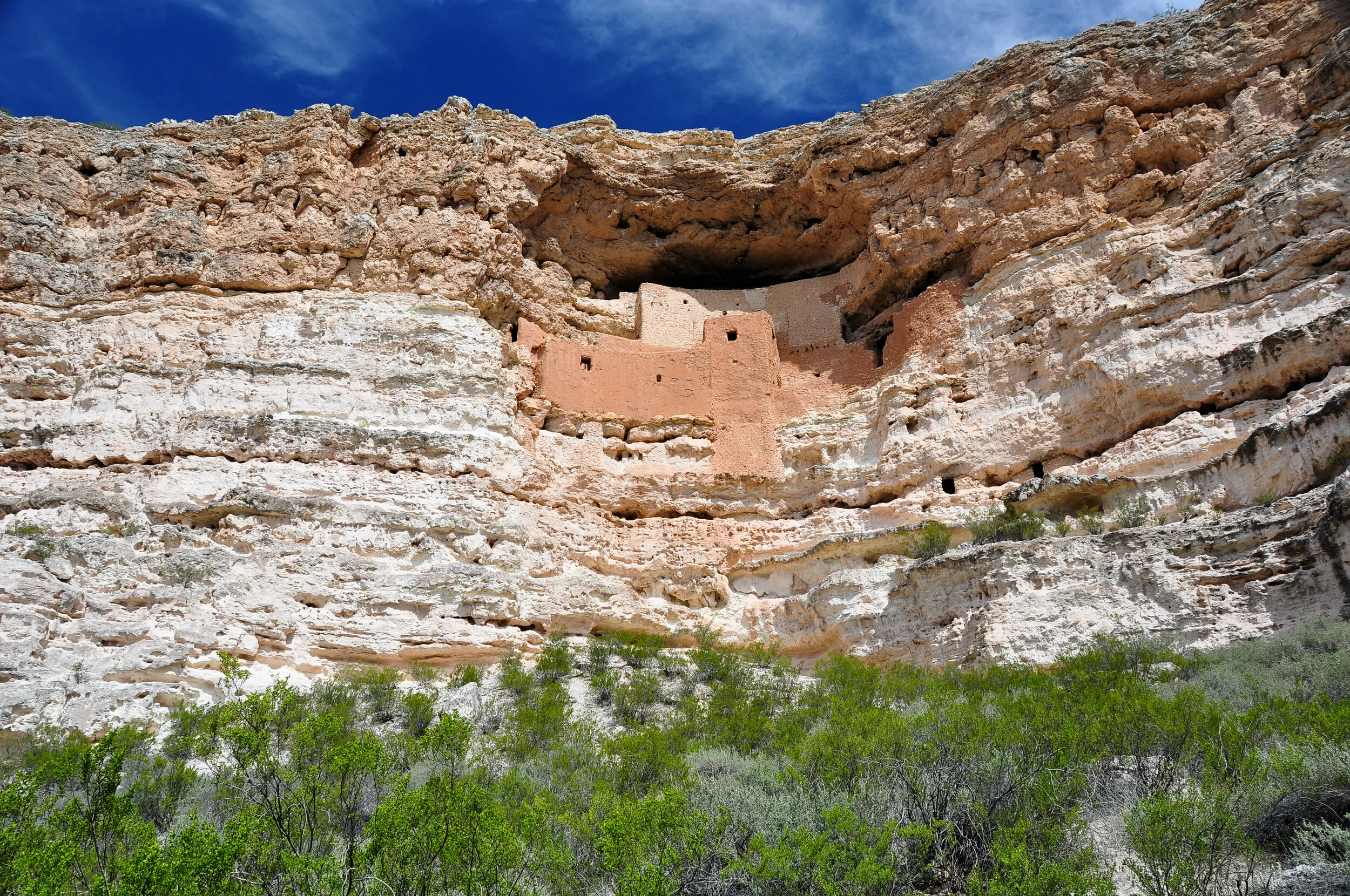 Ancient cliff dwelling built into a red and white layered rock formation with greenery at the base and a blue sky with some clouds above.