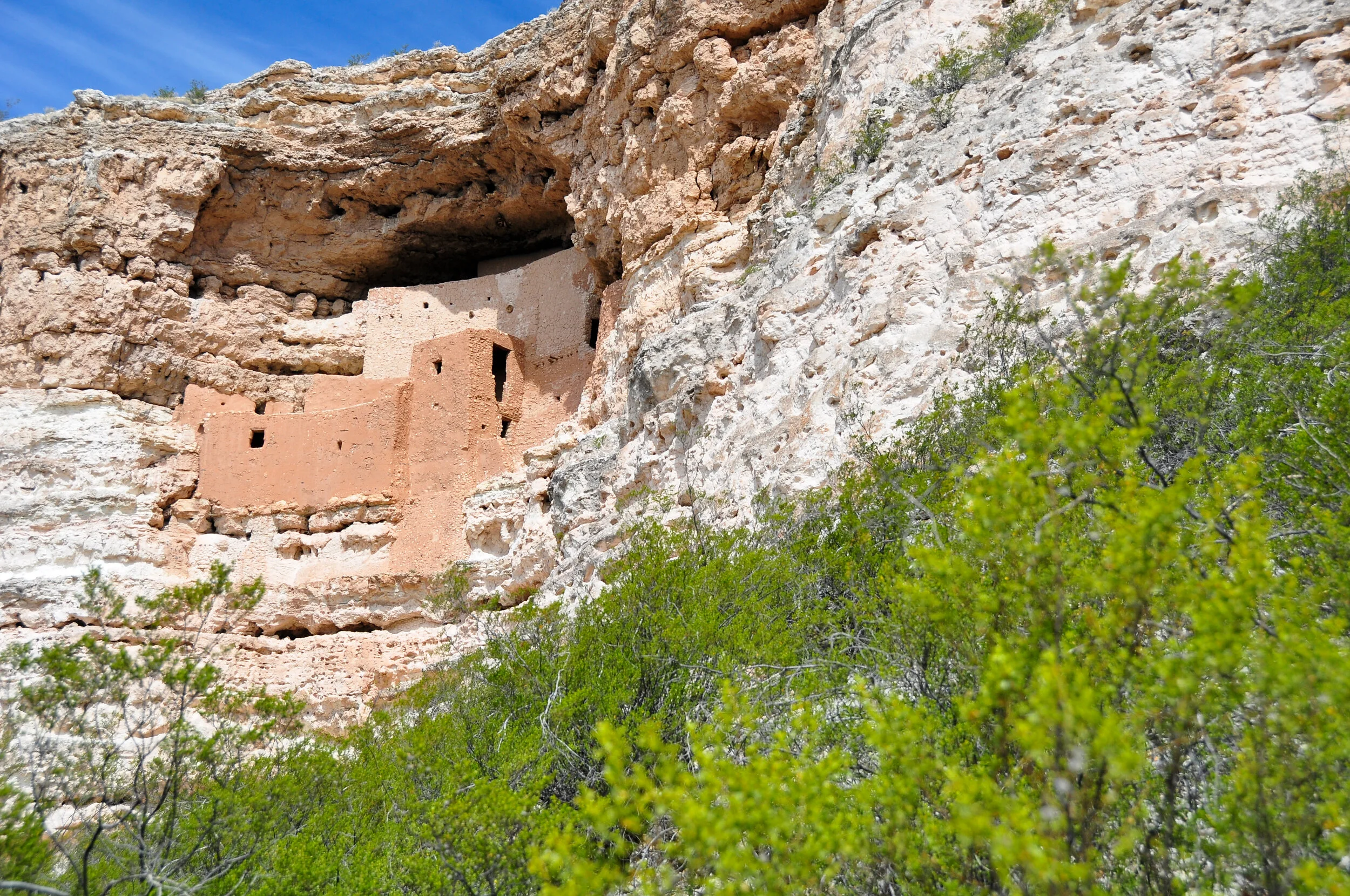 Ancient cliff dwelling built into a rock face, surrounded by green shrubbery, under a partly cloudy sky.