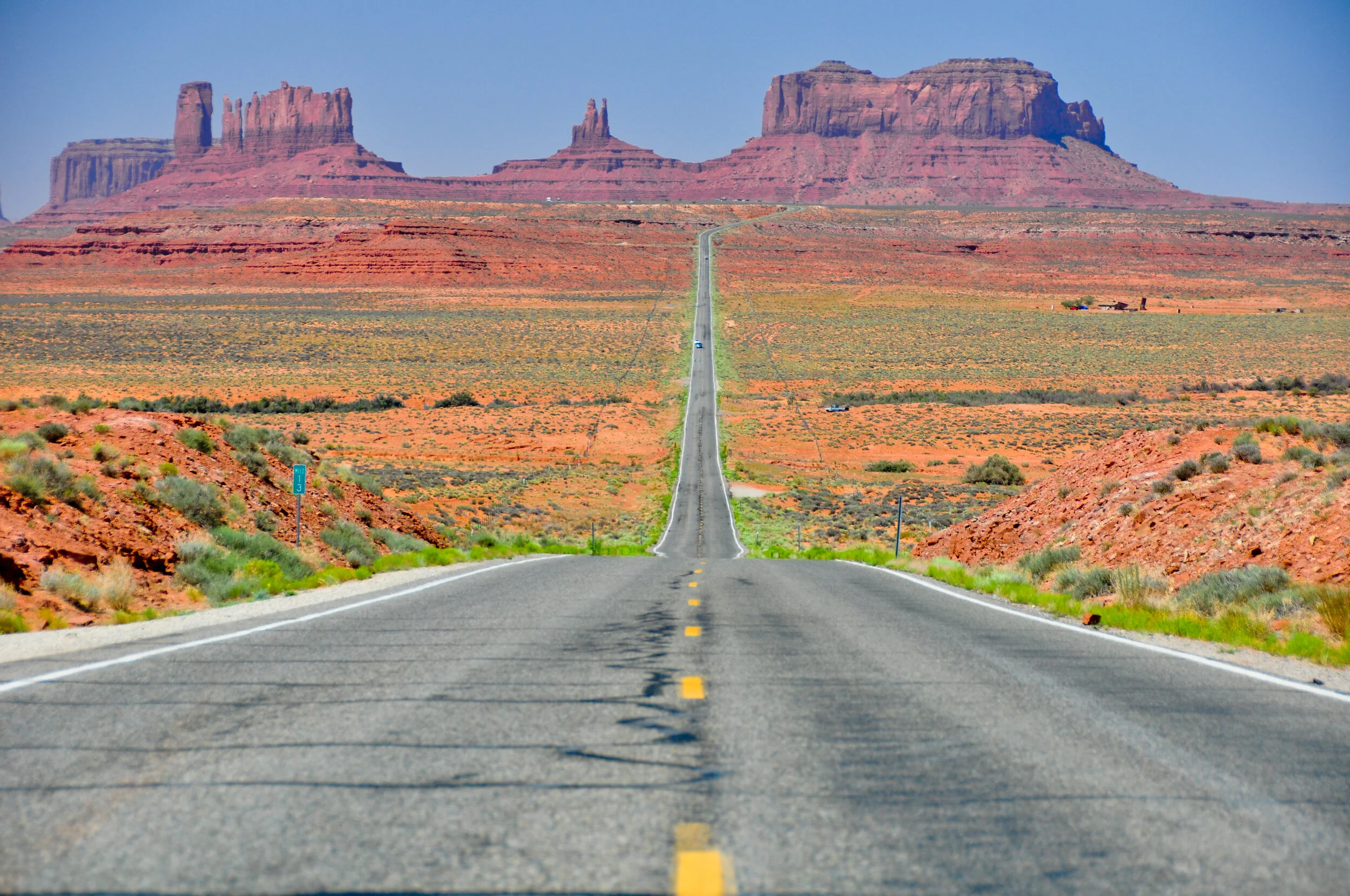 Long straight road cutting through a desert landscape with red rock formations and mesas in the distance under a clear blue sky.