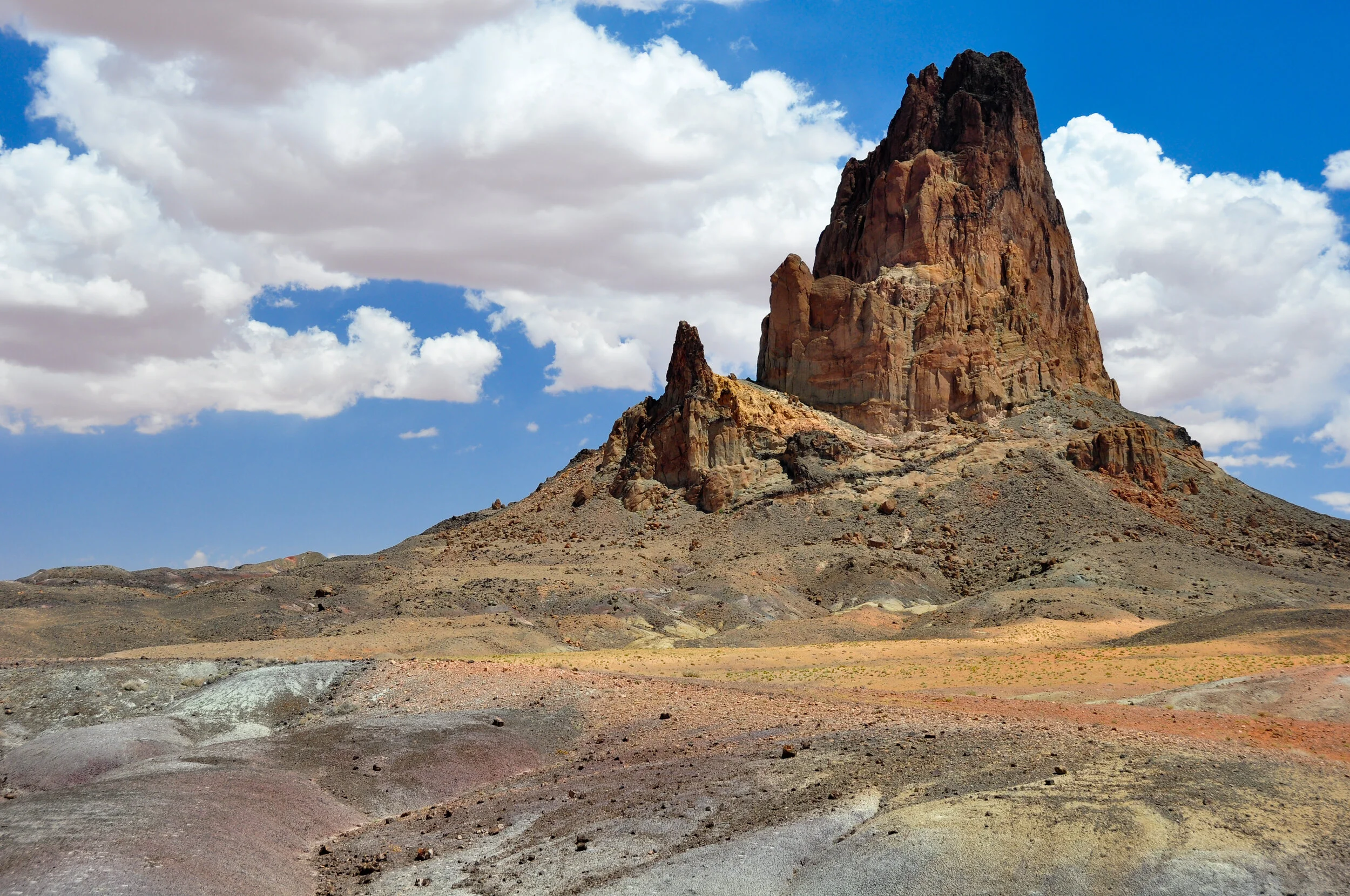 A large, rocky desert landscape featuring a prominent tall mountain or butte with a rugged, reddish-brown surface, under a partly cloudy sky.