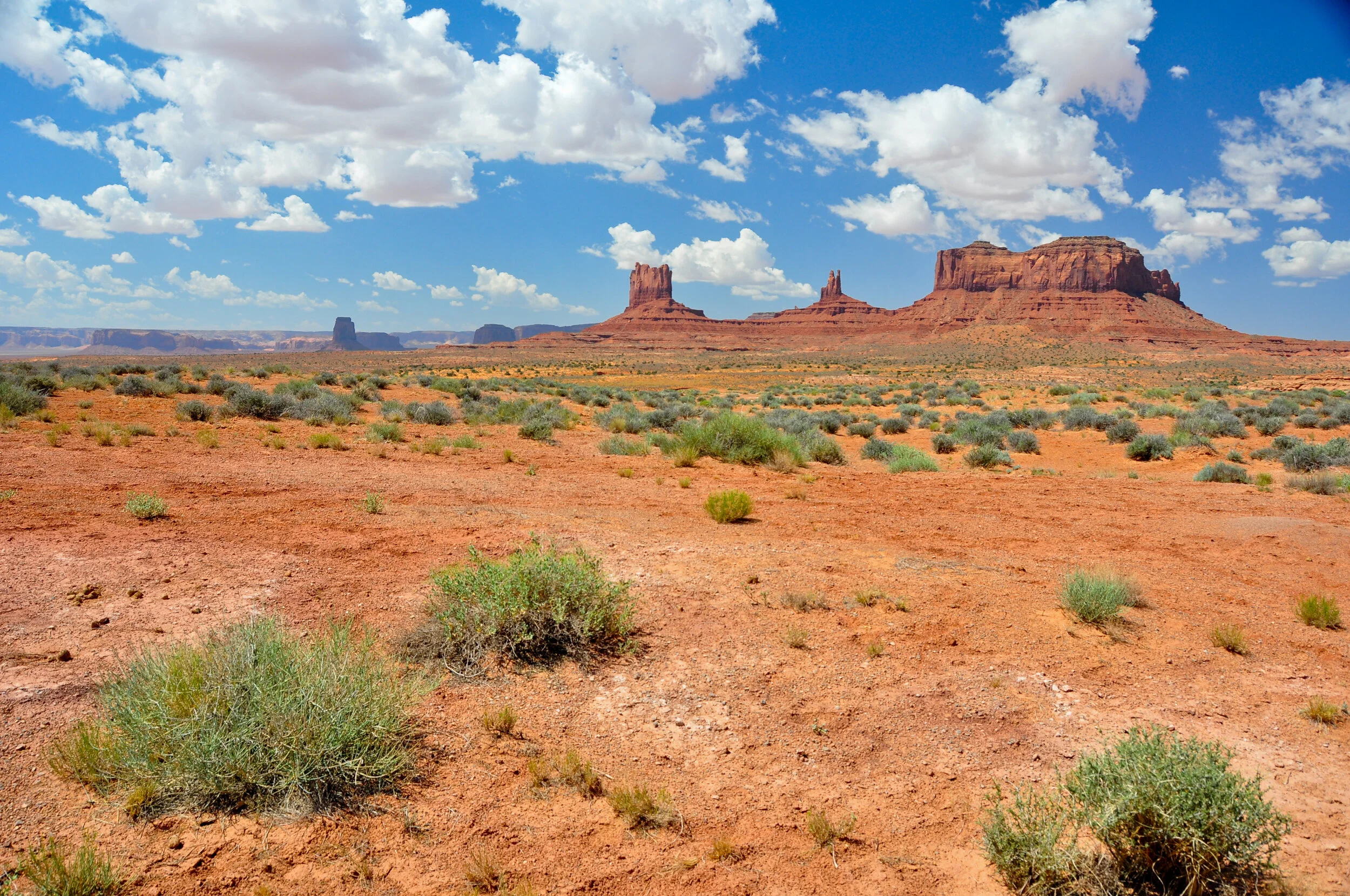 Desert landscape with red rock formations and sparse green bushes under a partly cloudy sky.