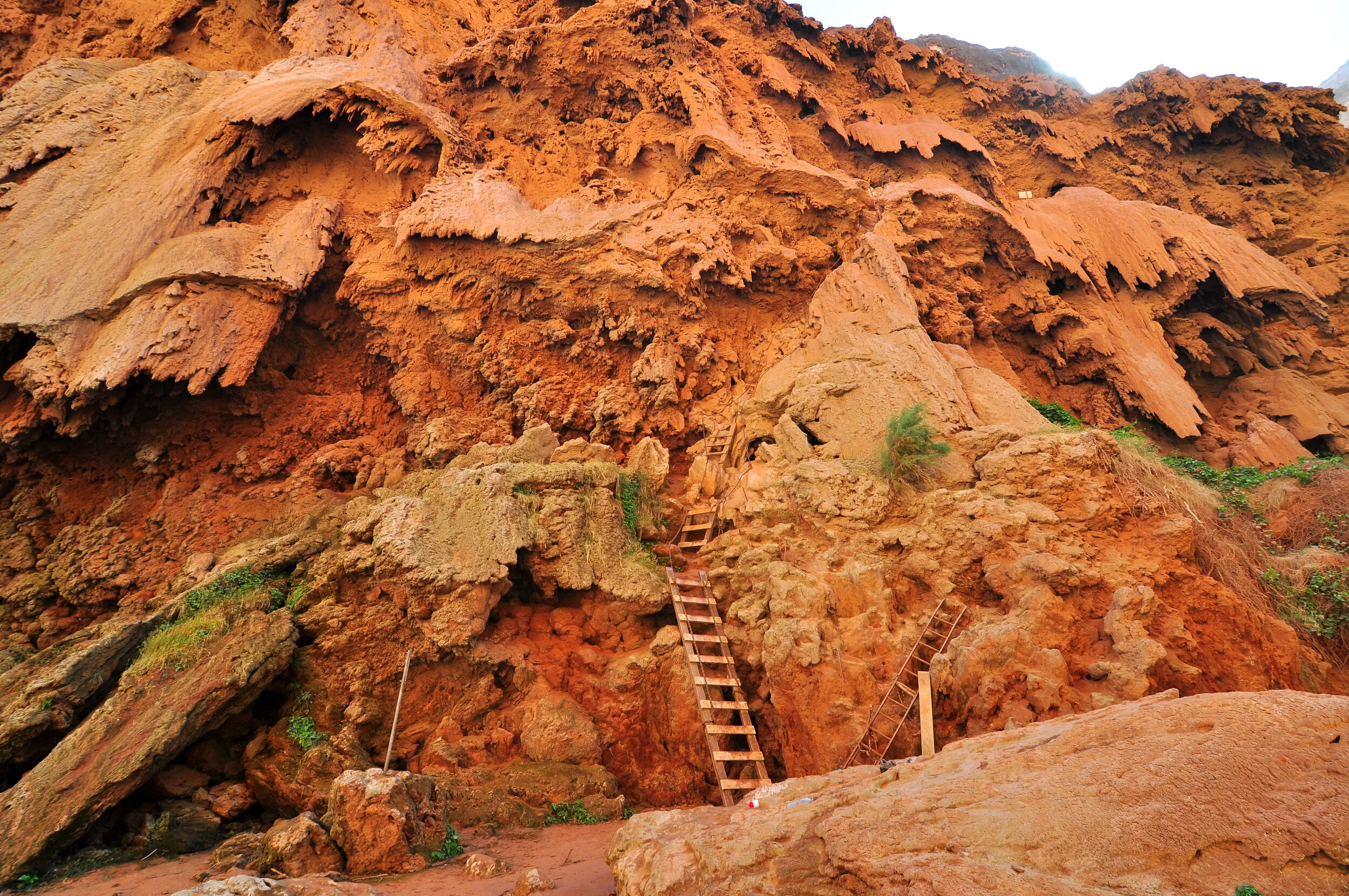 A rugged, reddish-brown rock cliff with wooden stairs and ladders leading up the face of the rock formation, with some green vegetation at the base and on the rock surface.