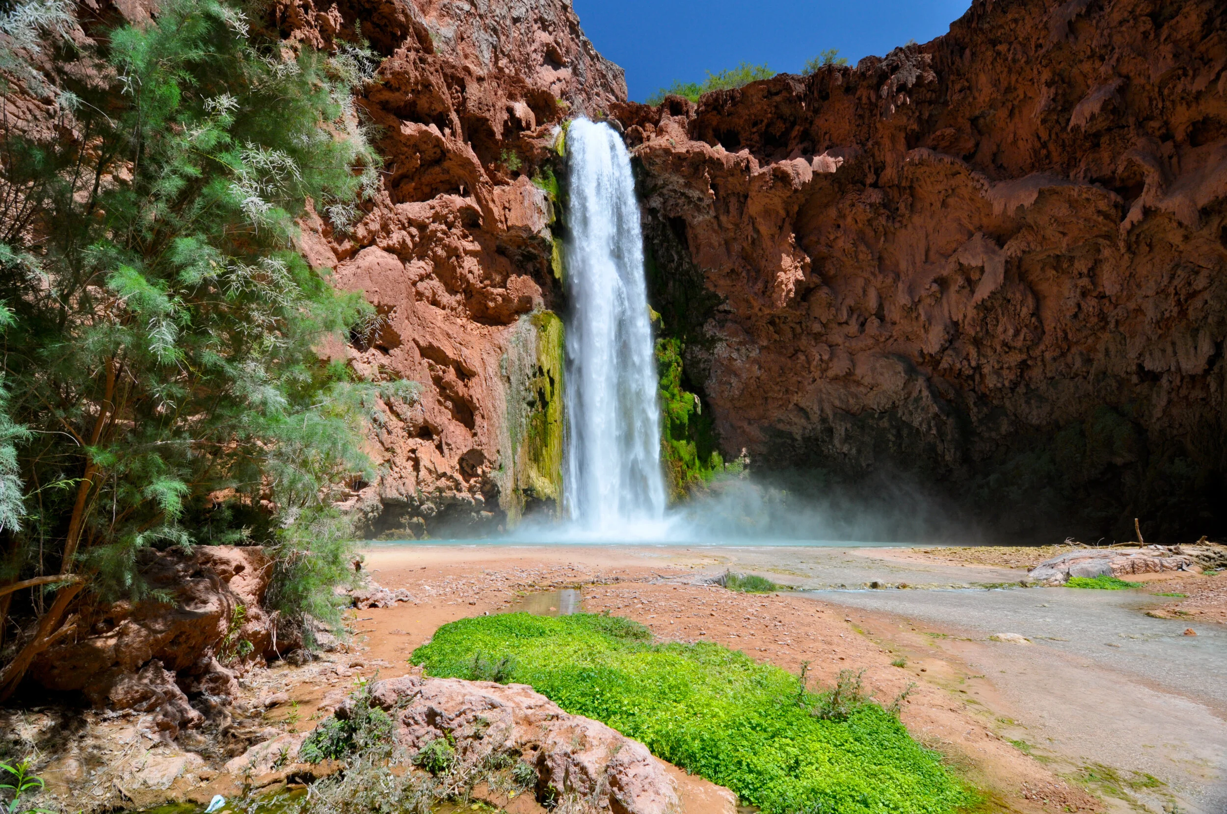 Waterfall cascading down a red rock cliff into a shallow pool surrounded by greenery, with a dirt trail in the foreground and a clear blue sky overhead.