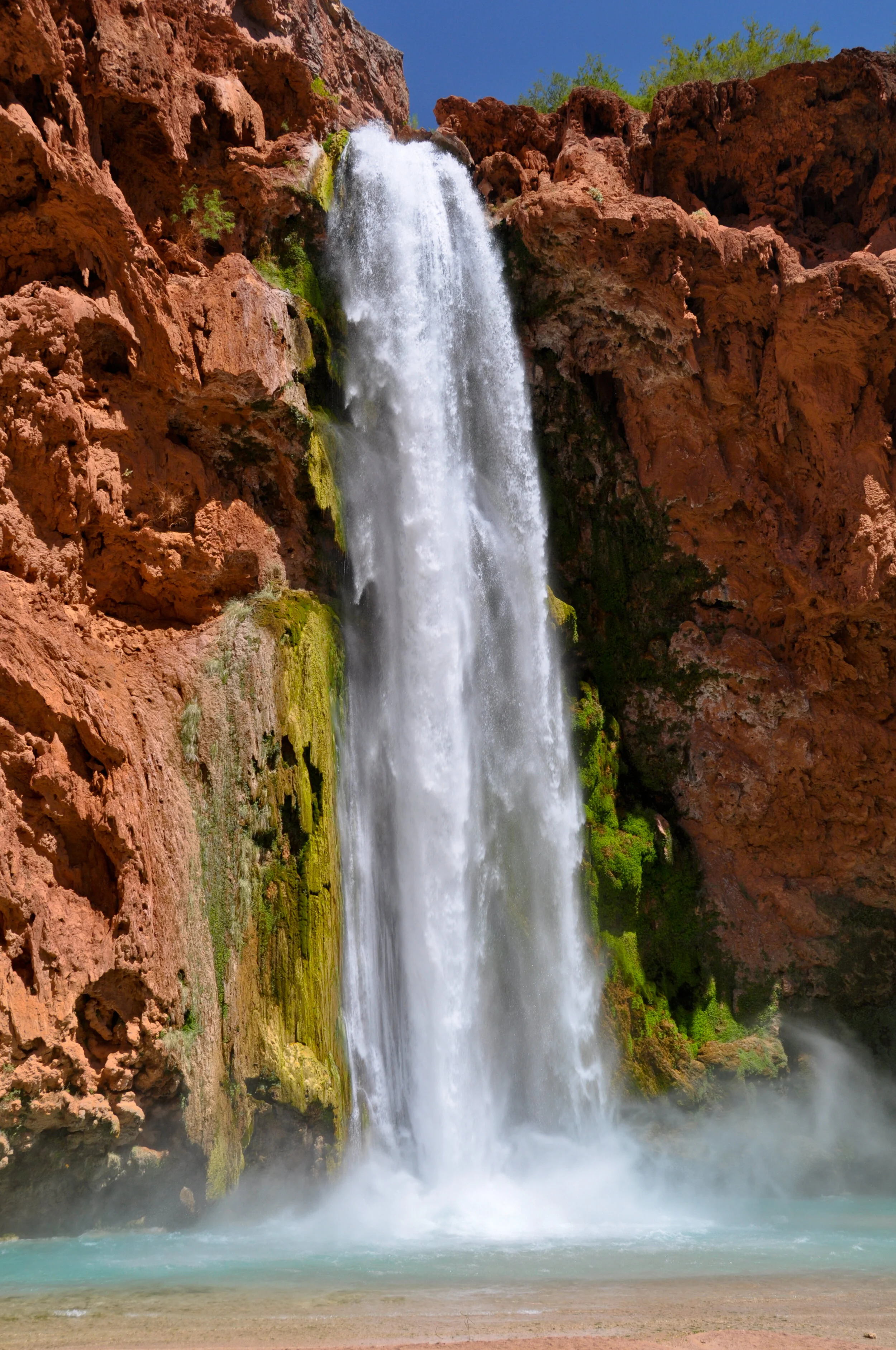 A tall waterfall cascading down red rock cliffs into a pool below, with some green moss and vegetation on the rocks.