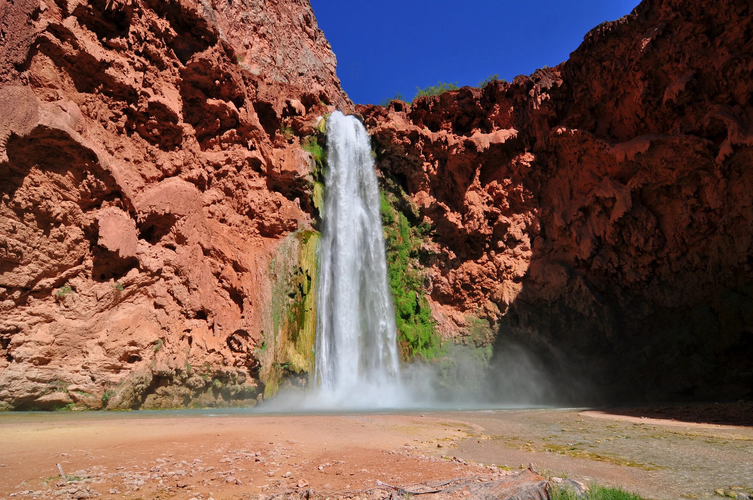 A tall waterfall cascading down red rocky cliffs into a shallow pool below, with green moss and plants on the rocks and a clear blue sky overhead