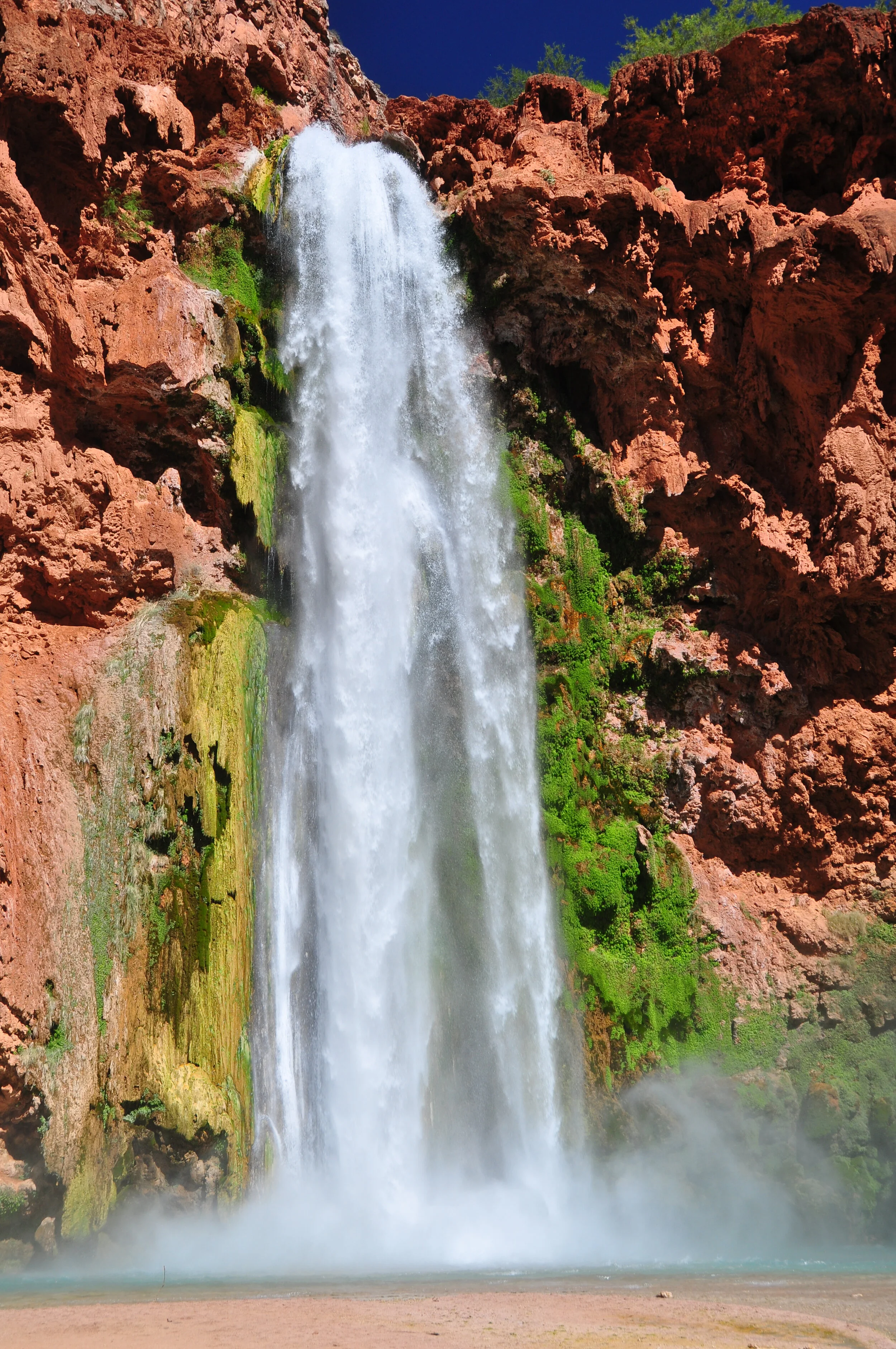 A tall, powerful waterfall cascading down red rocky cliffs into a misty pool below, with bright green vegetation around it under a clear blue sky.