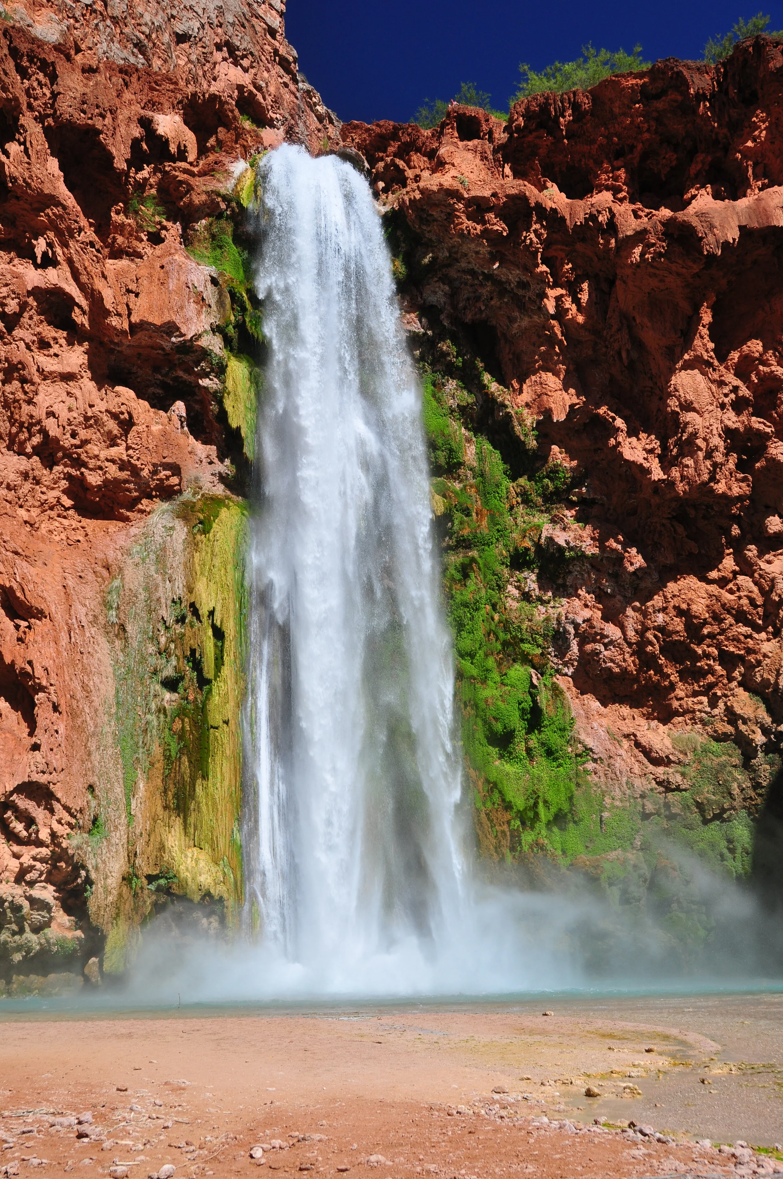 A waterfall flowing down a red rocky cliff with green moss and vegetation, with mist rising at the base and a clear blue sky overhead.
