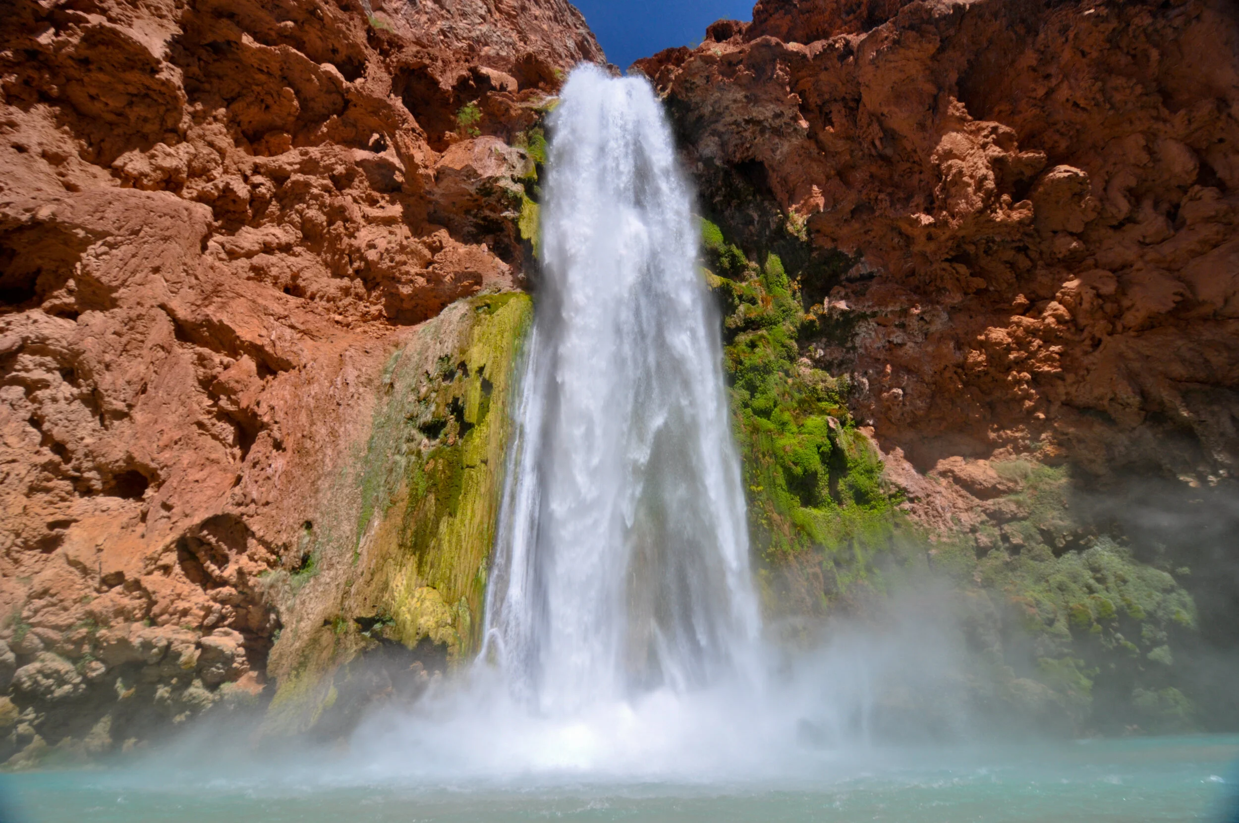 A large waterfall flowing over a rocky red canyon with green moss and foliage, mist rising from the water at the bottom.