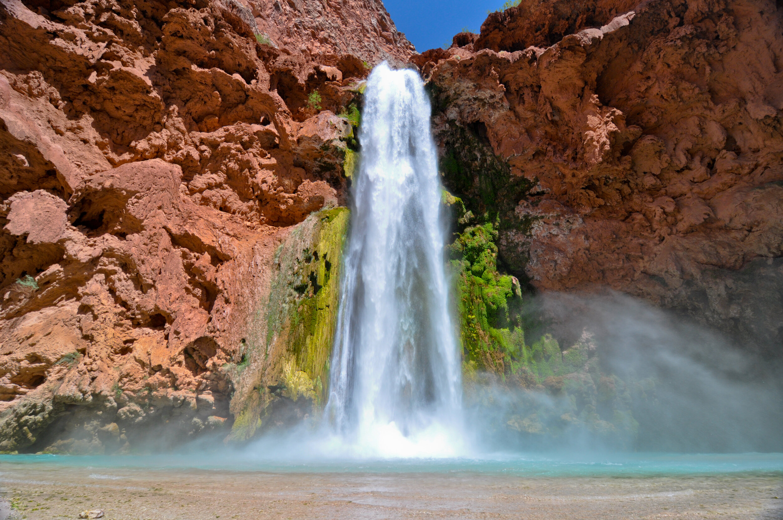 Waterfall flowing down red rock cliffs into a pool of water below, with green moss and plants on the rocks, and blue sky overhead.