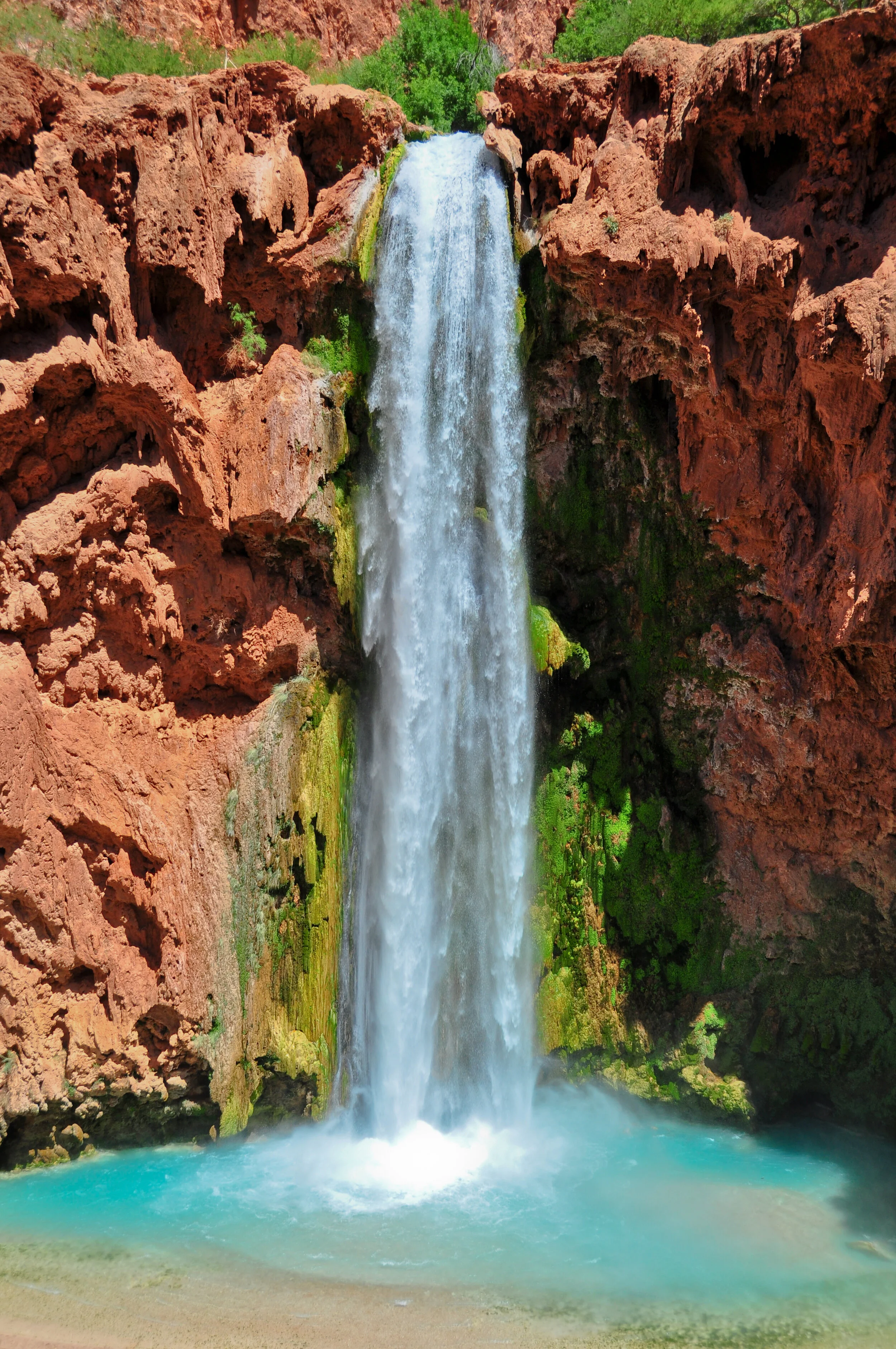 Waterfall flowing down from a rocky cliff into a blue pool below, surrounded by green vegetation and red rock formations.