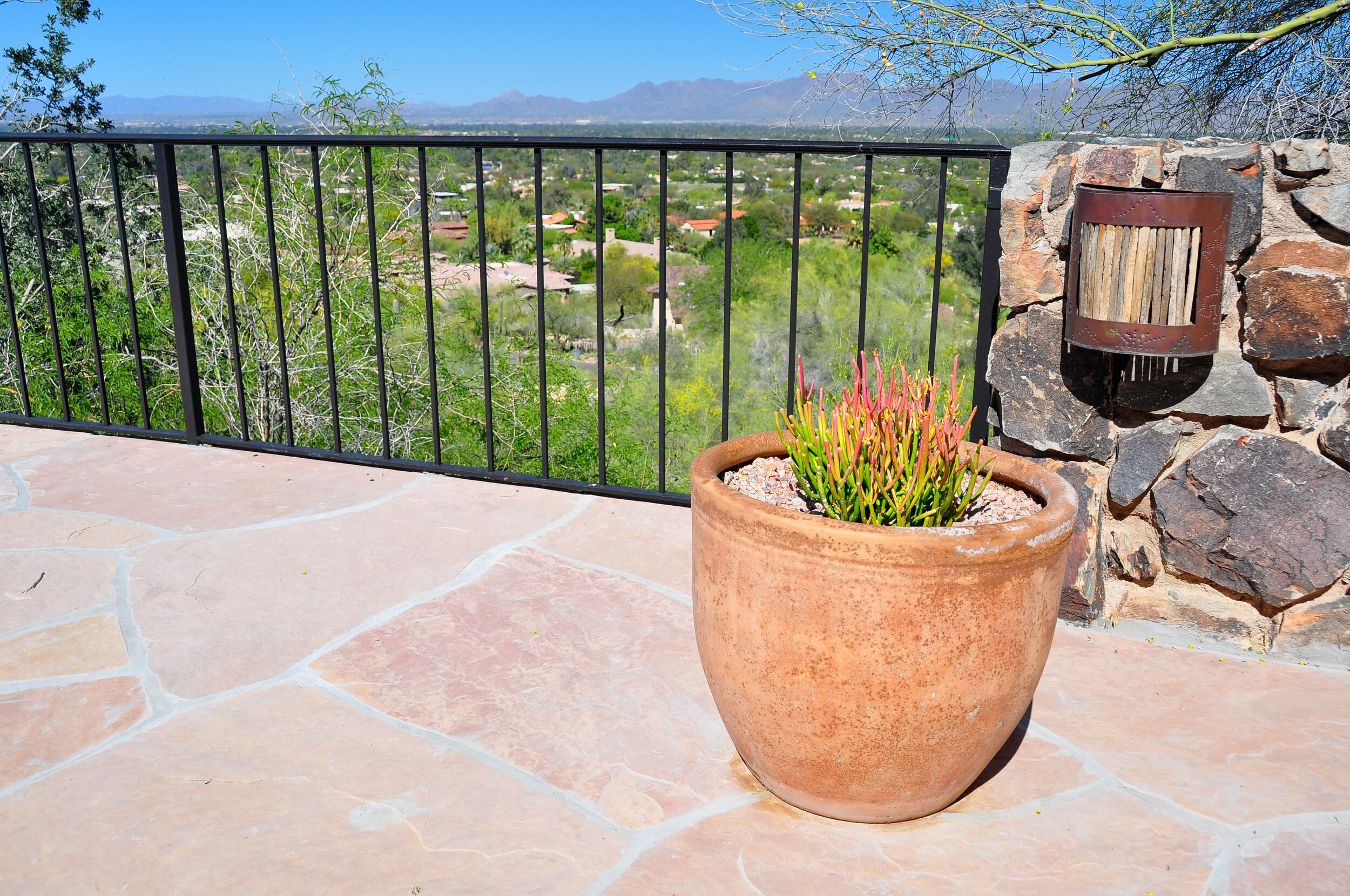Terrace with large beige potted succulent, stone wall with a wall-mounted mailbox, view of trees and houses in the distance, mountains on the horizon, clear blue sky.