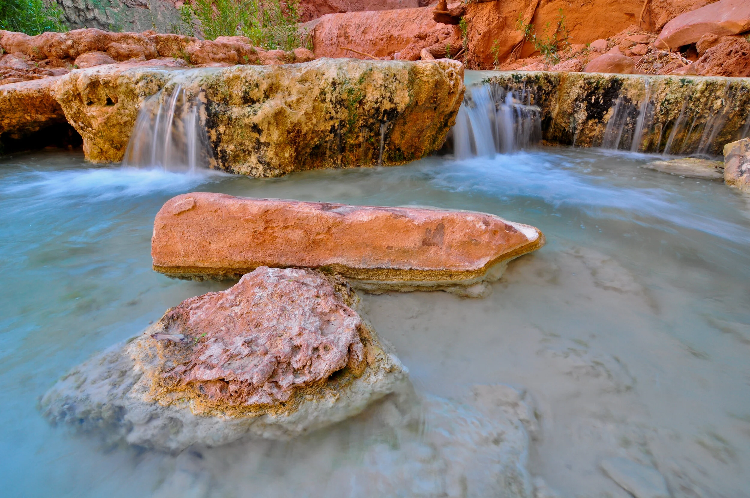 Small waterfall flowing over pink and brown rocks in a natural setting with surrounding scenic landscape.