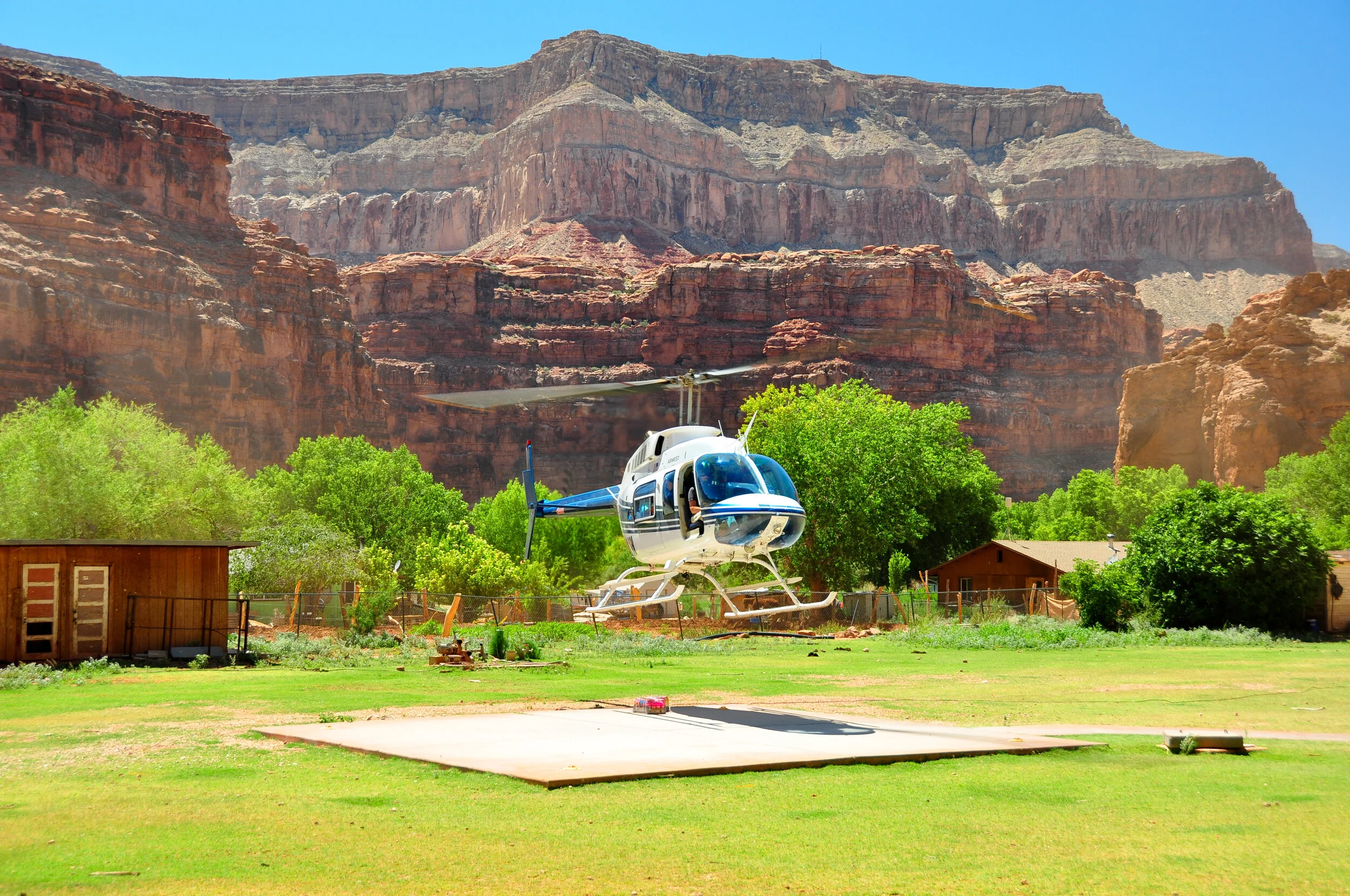 A helicopter hovering above a helipad with green trees and houses in the background, set against tall red canyon cliffs and a clear blue sky.