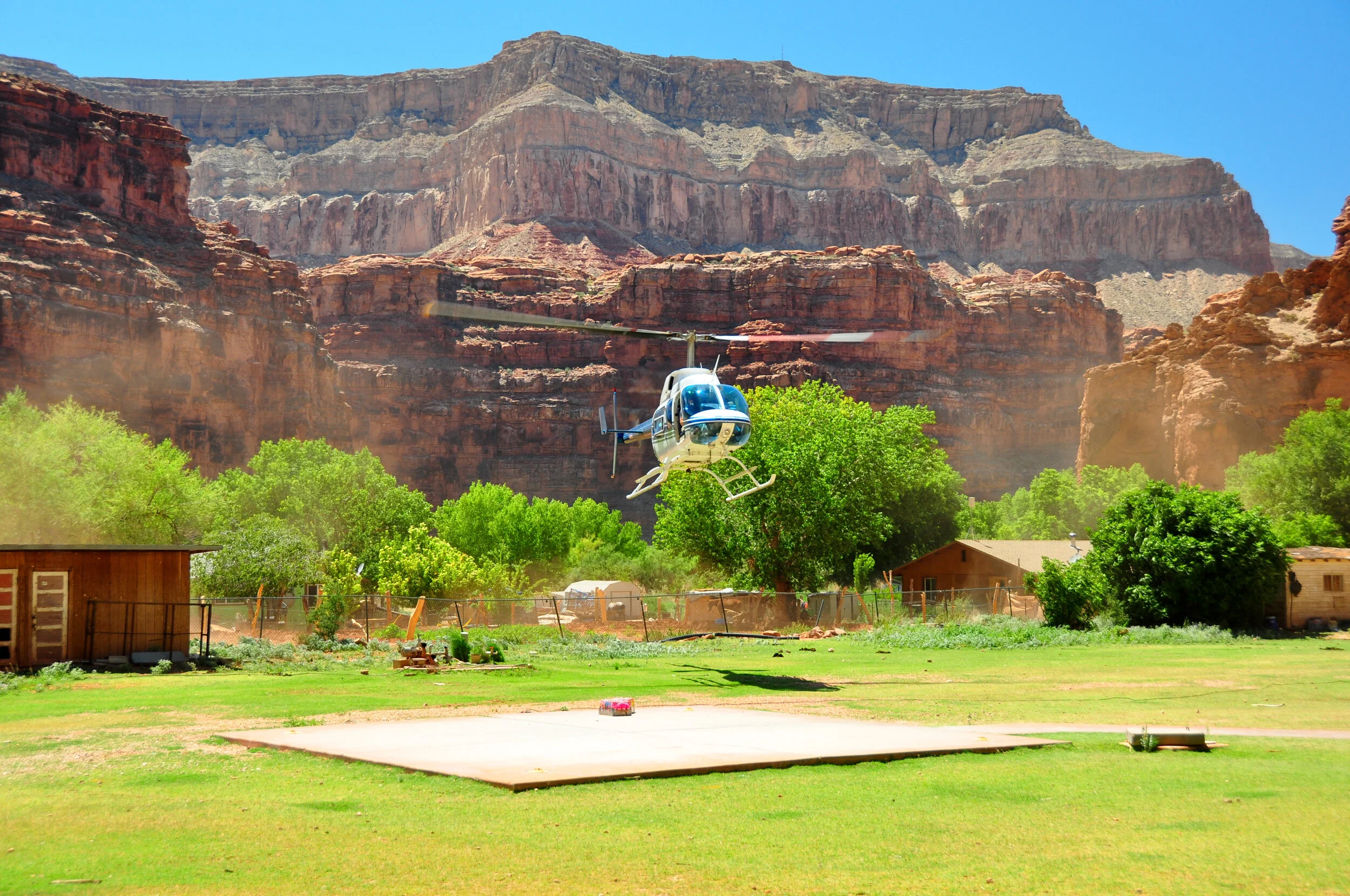 A helicopter flying low over a lush green yard with trees and small buildings, with a backdrop of towering red and brown canyon cliffs under a clear blue sky.