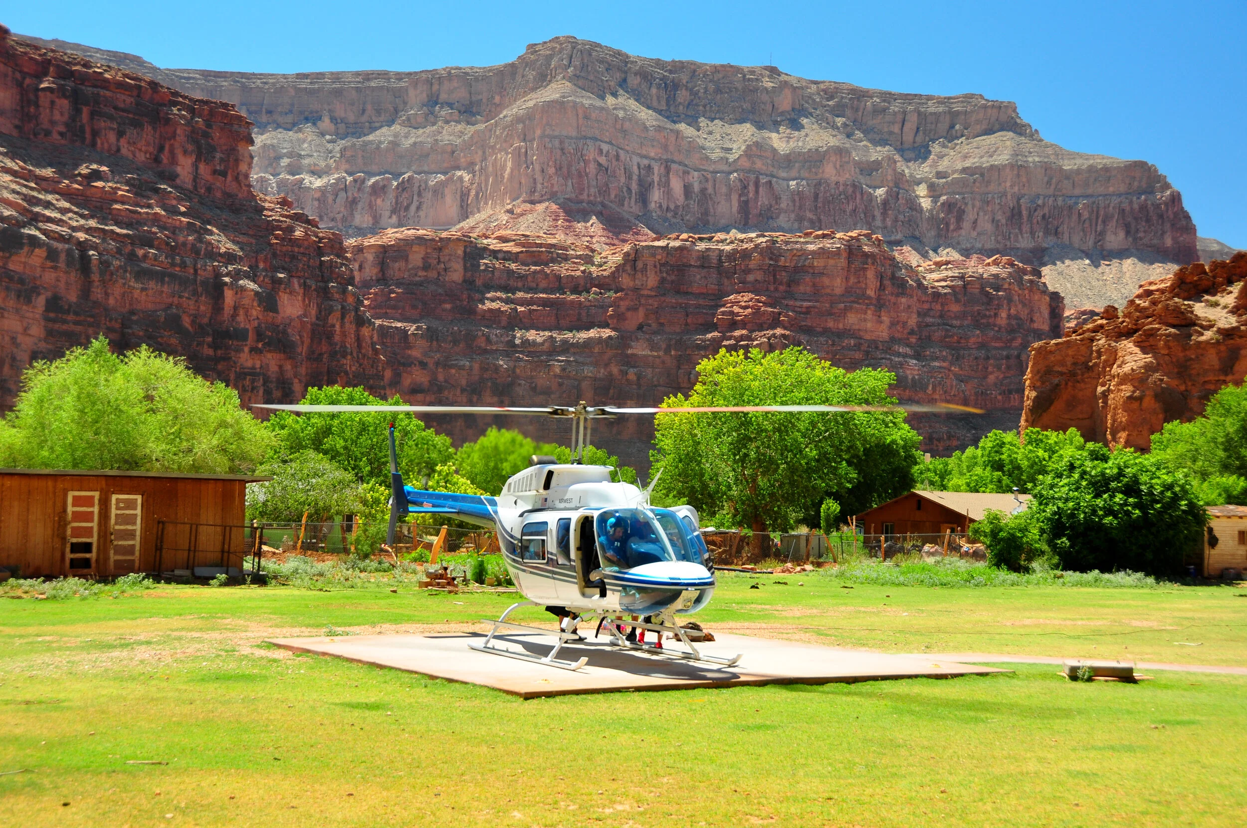 A white and blue helicopter parked on a concrete pad in a grassy area, with trees, small buildings, and red rock formations in the background under a clear blue sky.