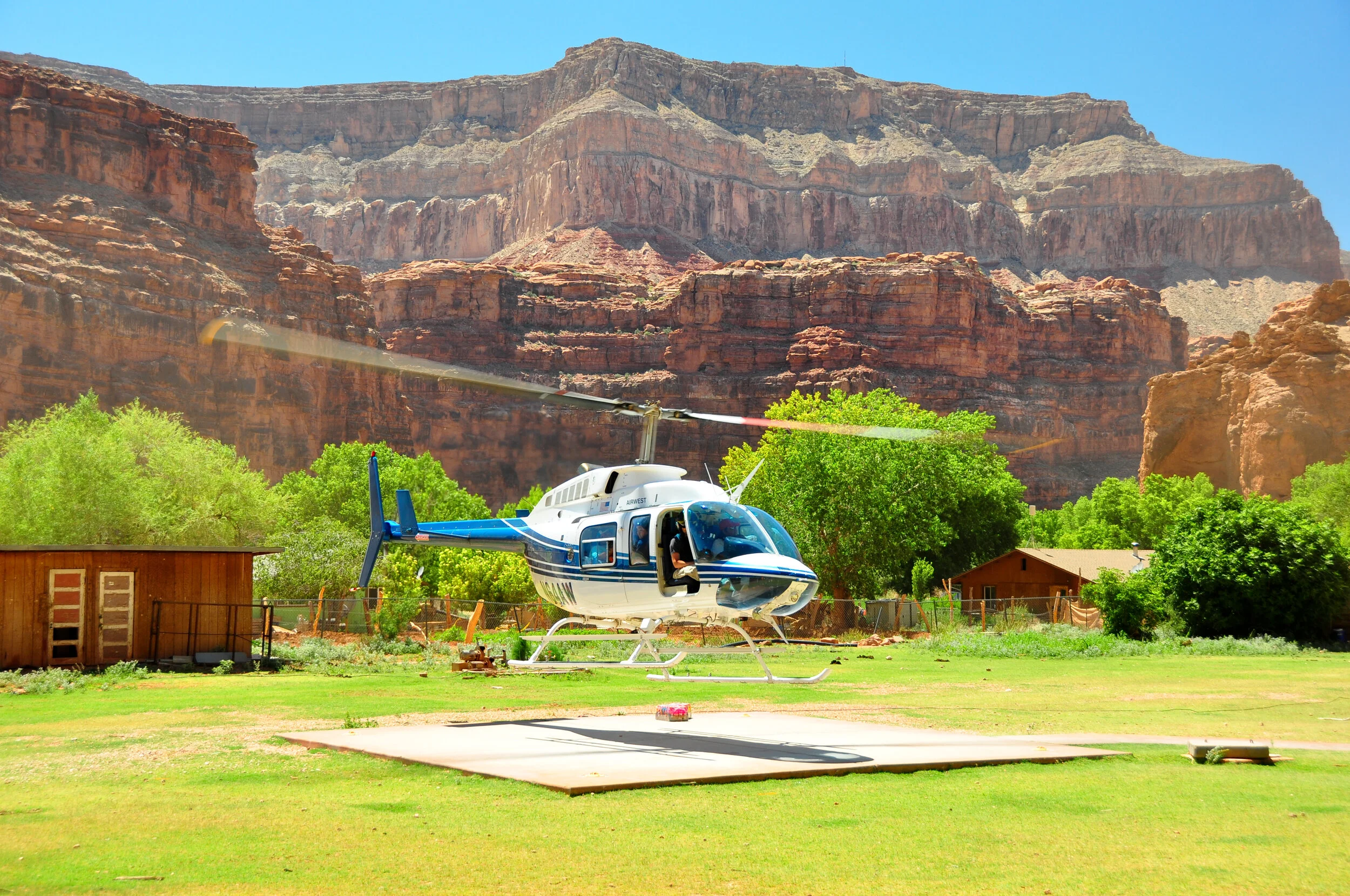 Helicopter parked on grassy field with red rock cliffs and blue sky in background, surrounded by trees and small buildings.