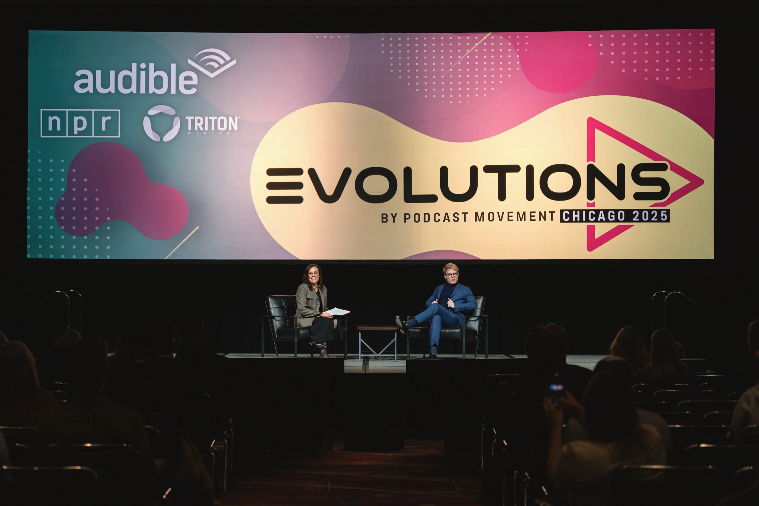 Two women seated on stage in front of a large screen displaying the event 'Evolutions by Podcast Movement Chicago 2025.' The screen features logos for Audible, NPR, Triton Digital, and has colorful abstract graphics.