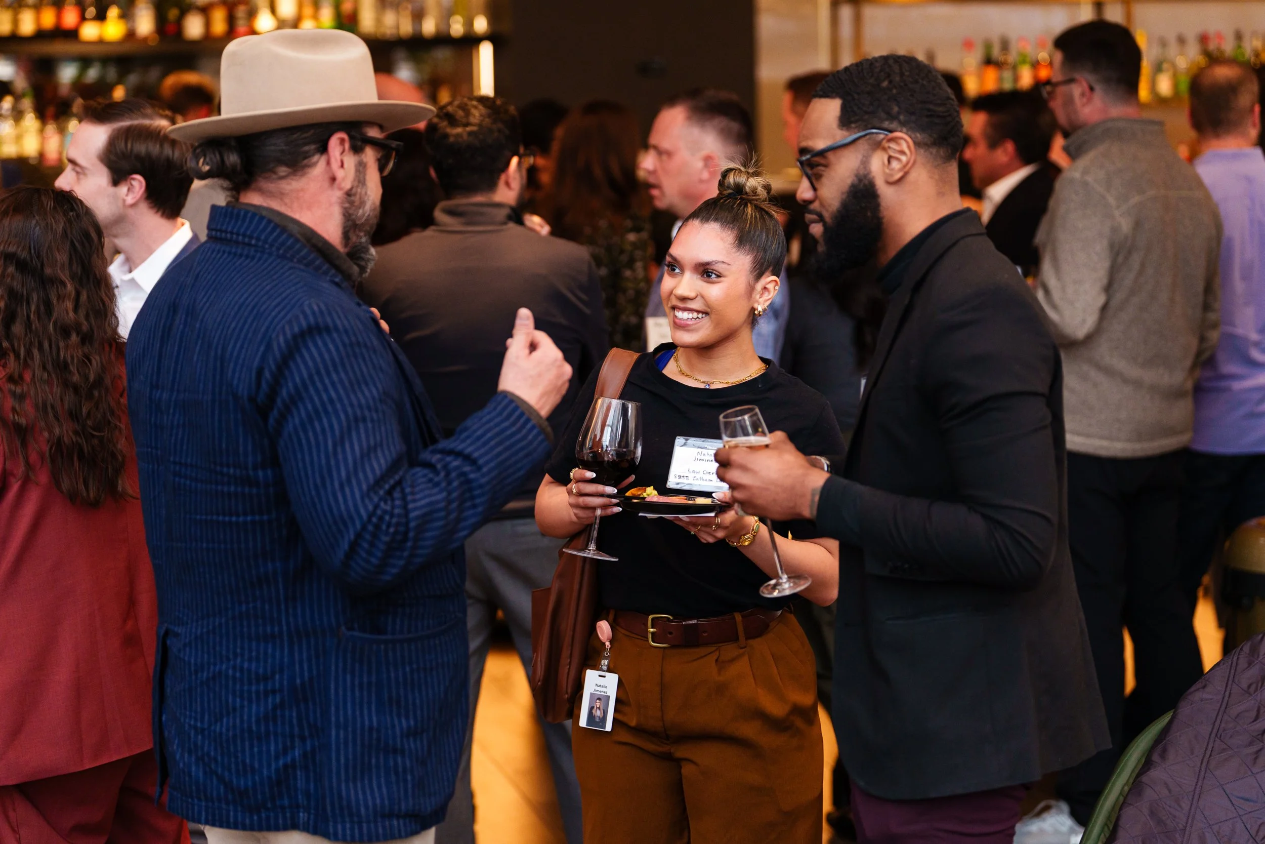 Three people are engaged in conversation at a social event in a bar or restaurant. The woman is smiling, holding a glass of red wine, and an appetizer plate. The man on the right is holding a wine glass, and the man on the left is wearing a hat and g