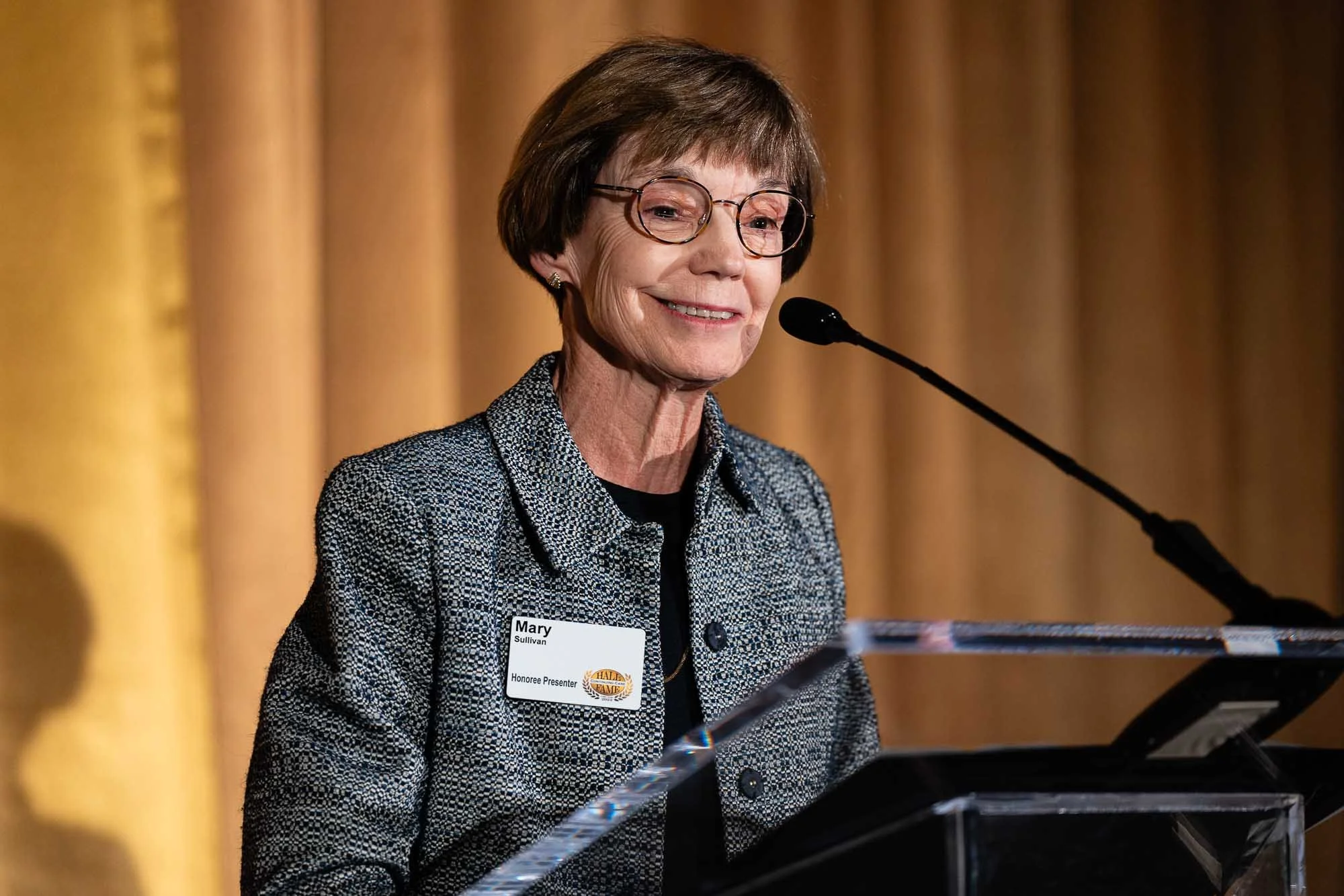 A woman with glasses and short brown hair speaking at a podium in front of a gold curtain, wearing a patterned blazer and a name tag that reads "Mary Sullivan, Honoree Presenter."