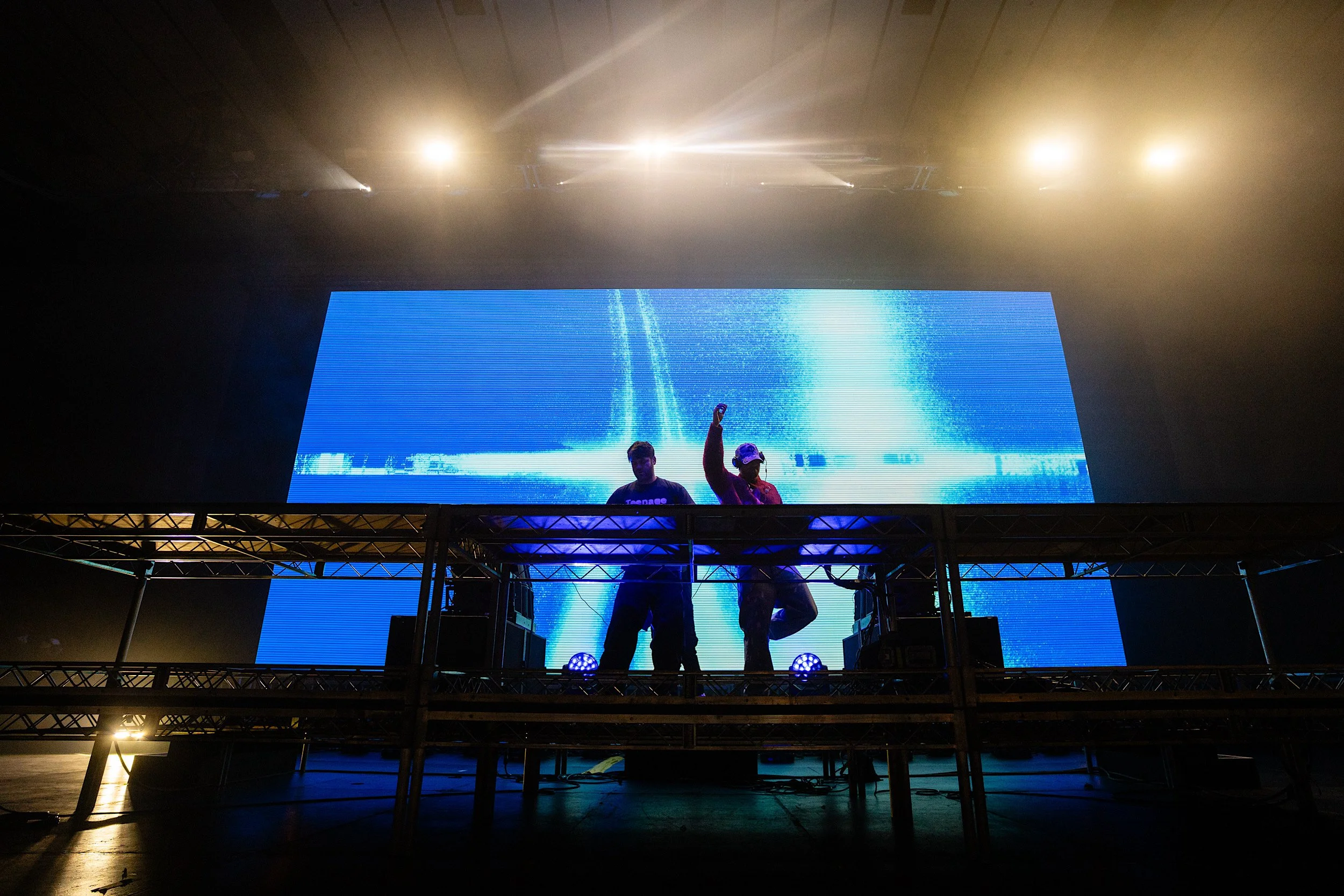 Two people on a DJ stage with a digital screen behind them displaying blue visuals, with stage lights overhead.