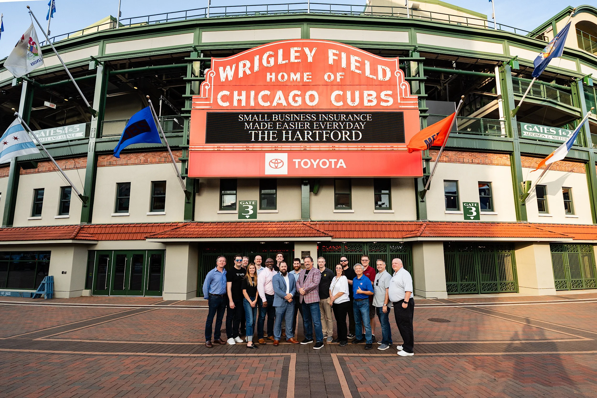 A group of people standing in front of Wrigley Field, home of the Chicago Cubs, on a brick walkway. The stadium features a large sign with advertisements and flags near the entrance.