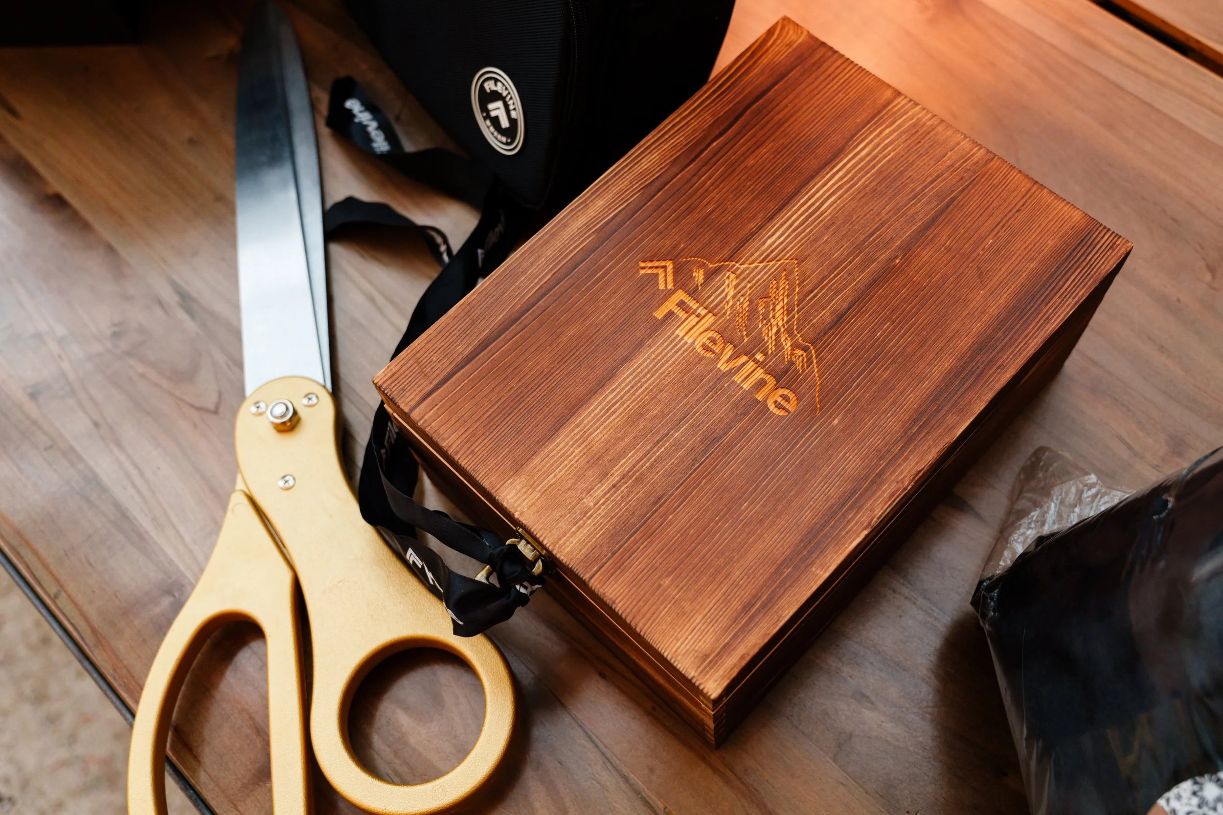 A wooden box with the word 'Traverse' and a mountain logo on the lid, a pair of large yellow scissors, and a black bag on a wooden table.