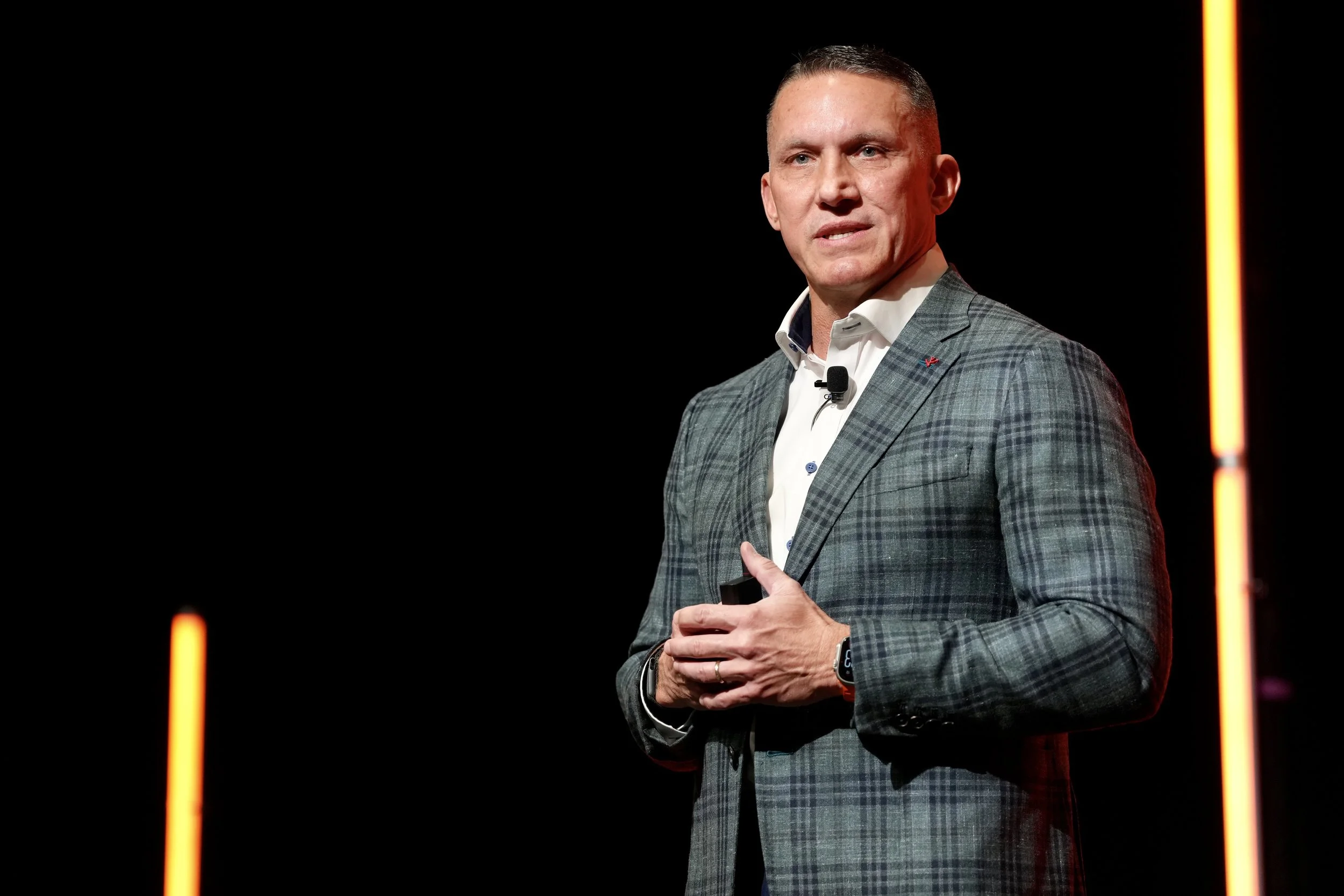 A man speaking on stage, wearing a gray plaid suit jacket, white shirt, and black wristwatch, with a black background and vertical orange lights.