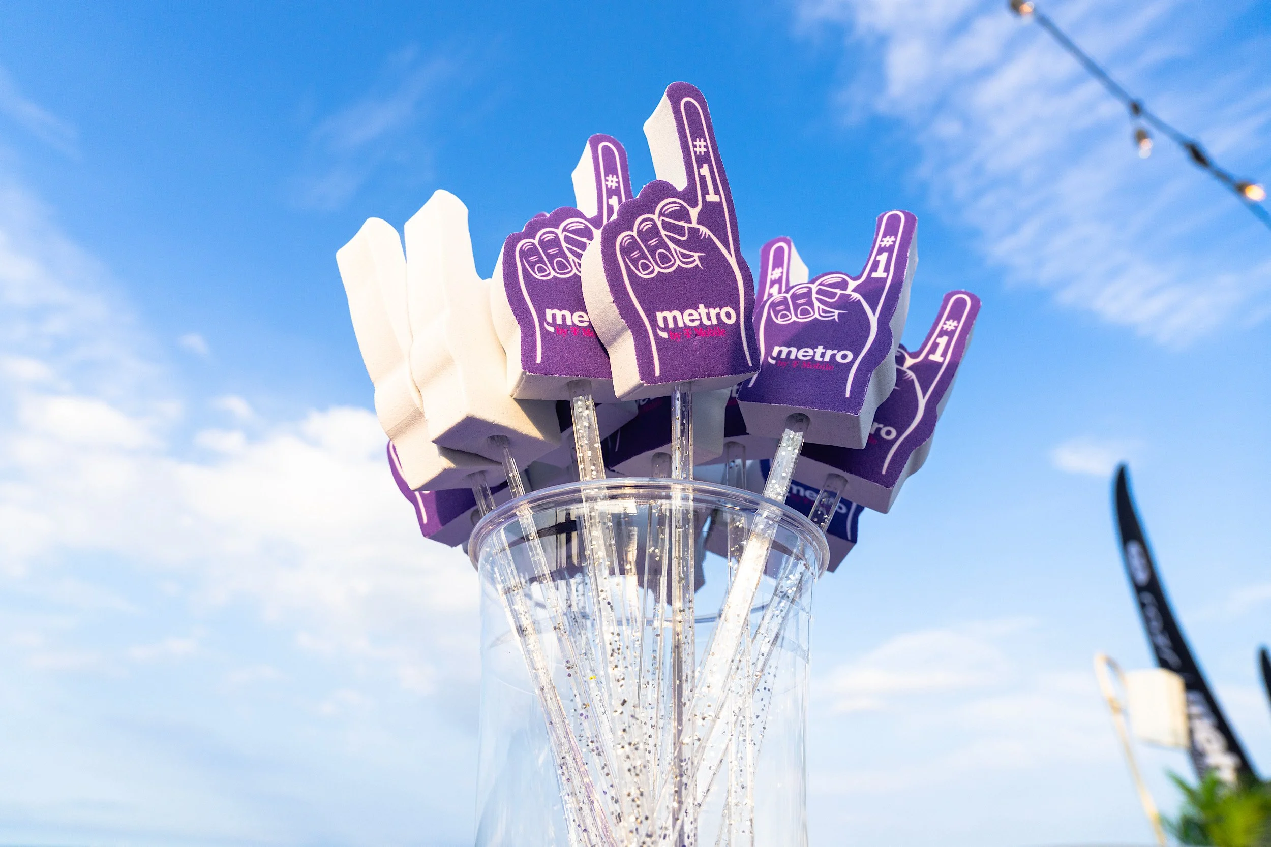 A clear glass jar filled with purple foam hand props with the word 'metro' and the number '#1' printed on them, set against a bright blue sky with scattered clouds.