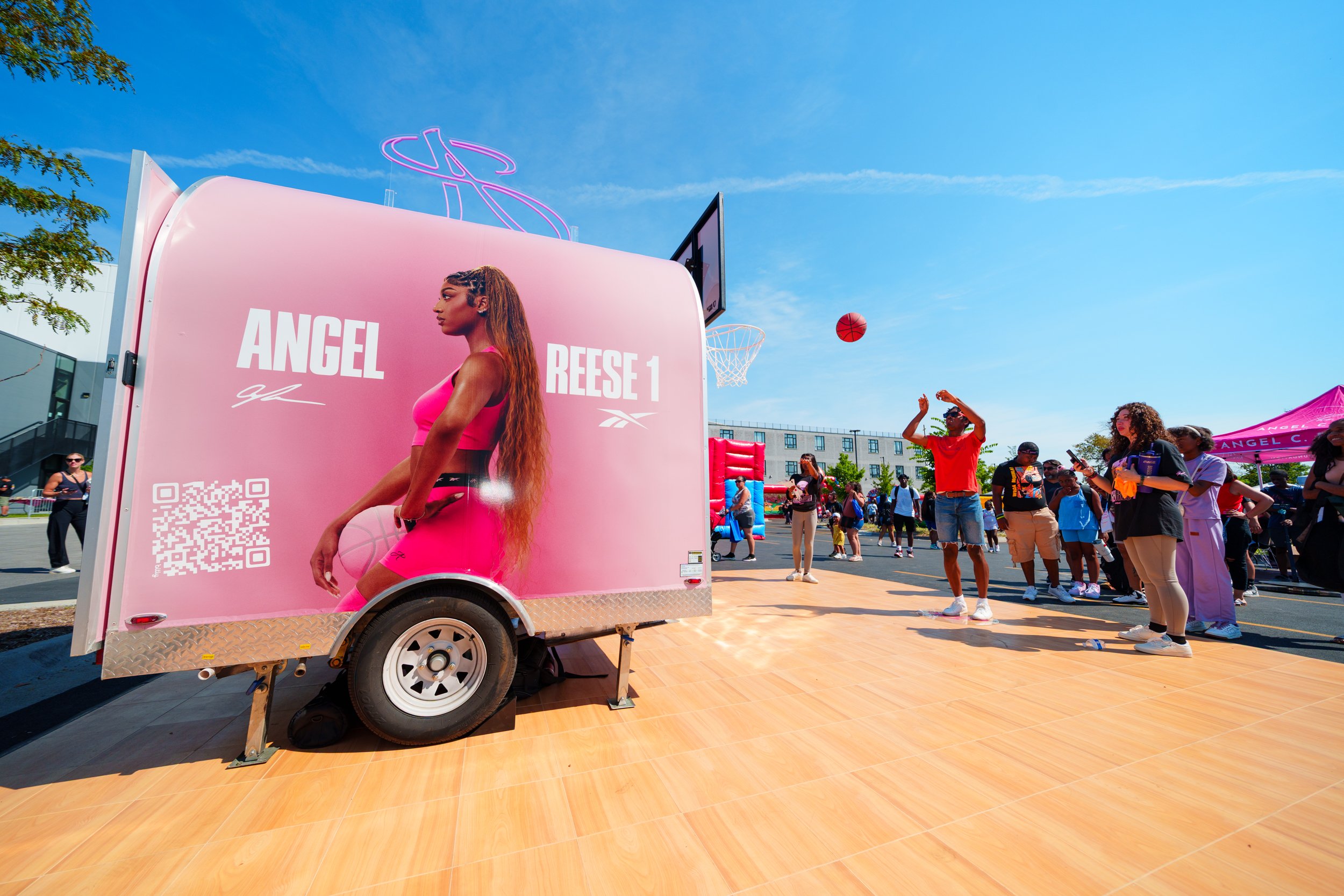 A mobile promotional trailer with a large image of a woman holding a basketball, wearing pink sports attire, and the text 'Angel Reese 1'. Several people are gathered around on a sunny day with a clear blue sky, some are watching a basketball shootin