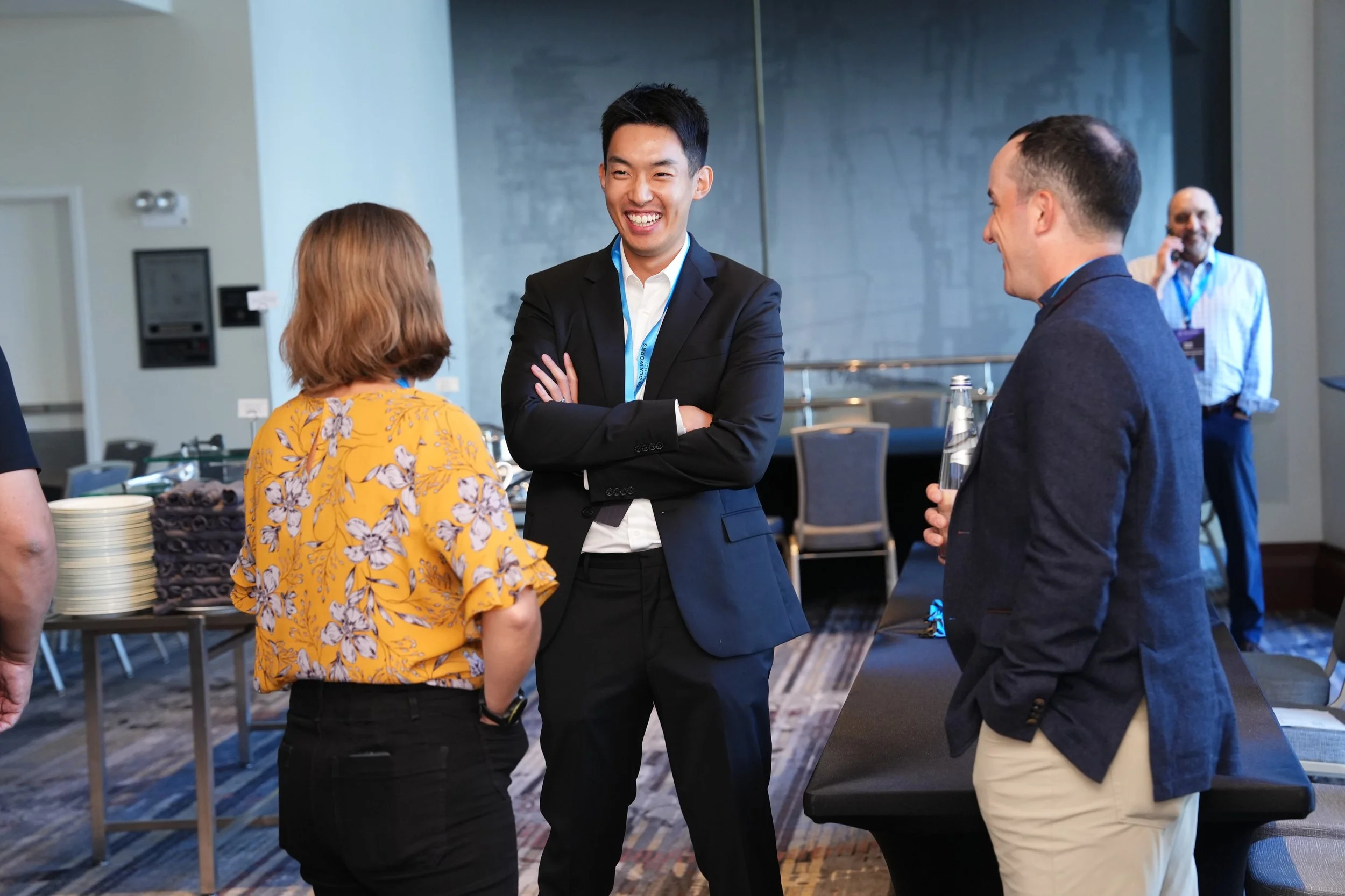 Four people engaging in conversation at a professional event or conference, with two men and a woman in the foreground and a man in the background talking on a cellphone.