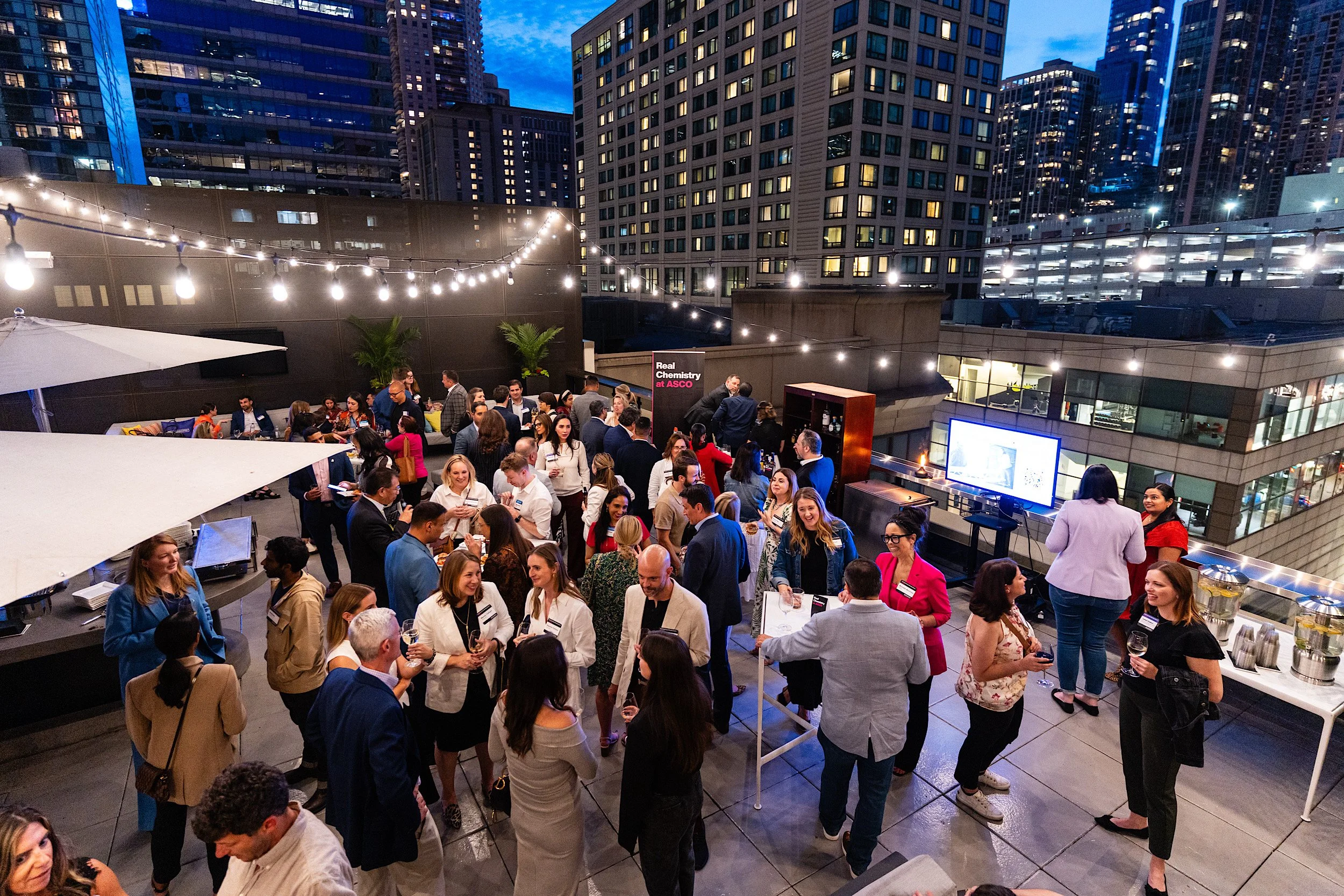 A rooftop cocktail party with people socializing in a city skyline setting during evening, with string lights, a large screen, and city skyscrapers in the background.