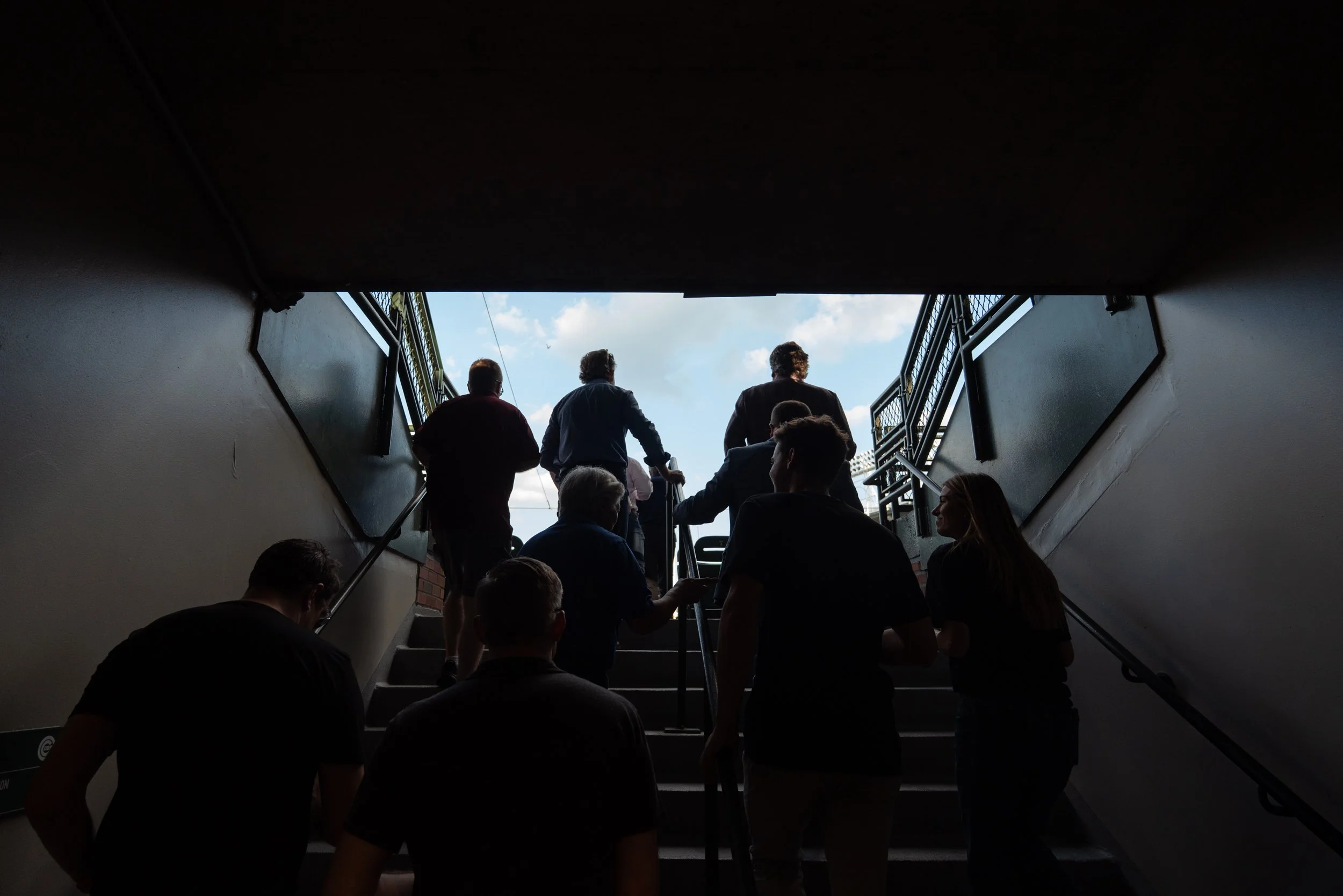 People walking up stairs toward a bright sky with clouds, viewed from underneath an overpass or tunnel.