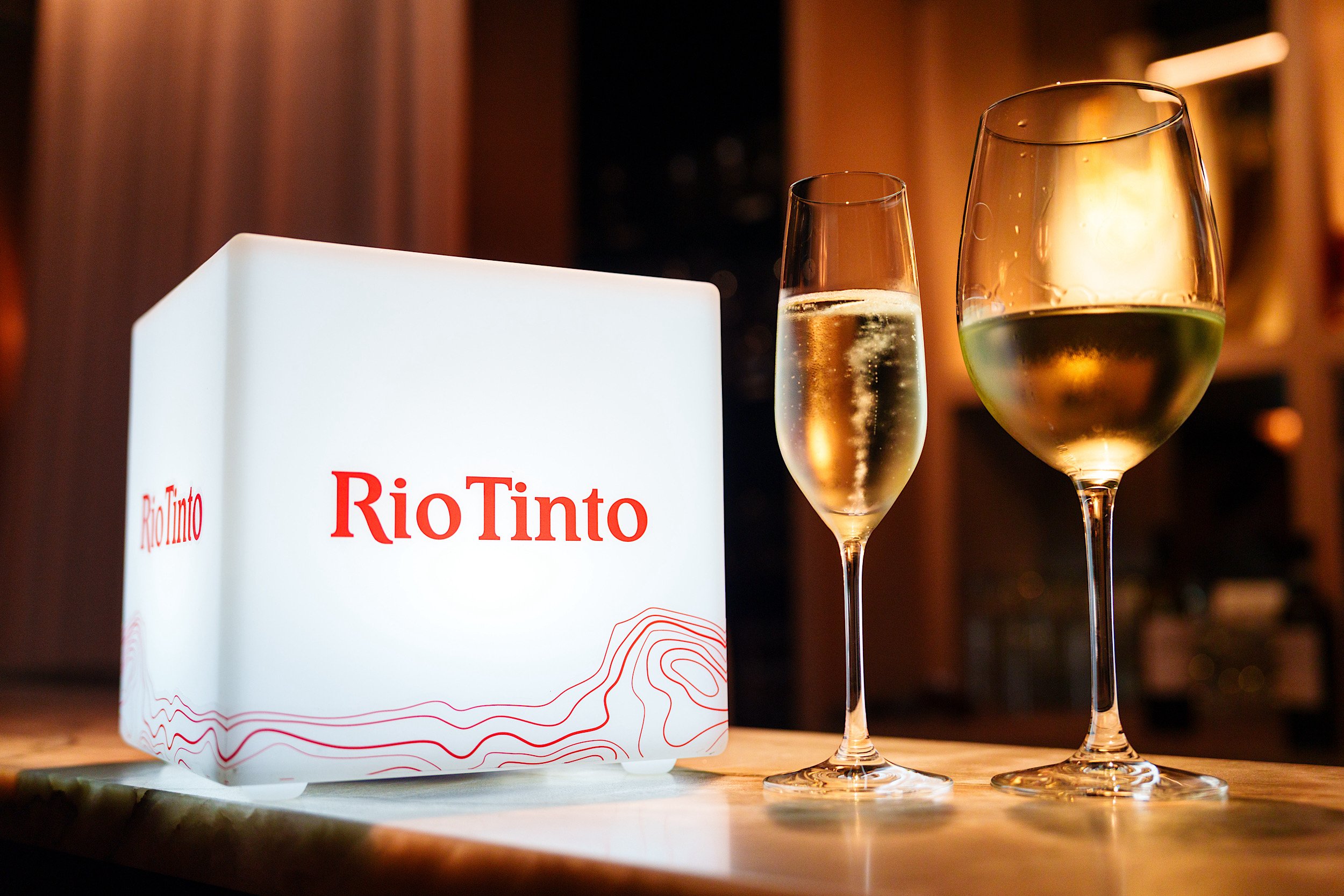 Two glasses of white wine and a box labeled 'Rio Tinto' on a wooden surface in a dimly lit room.