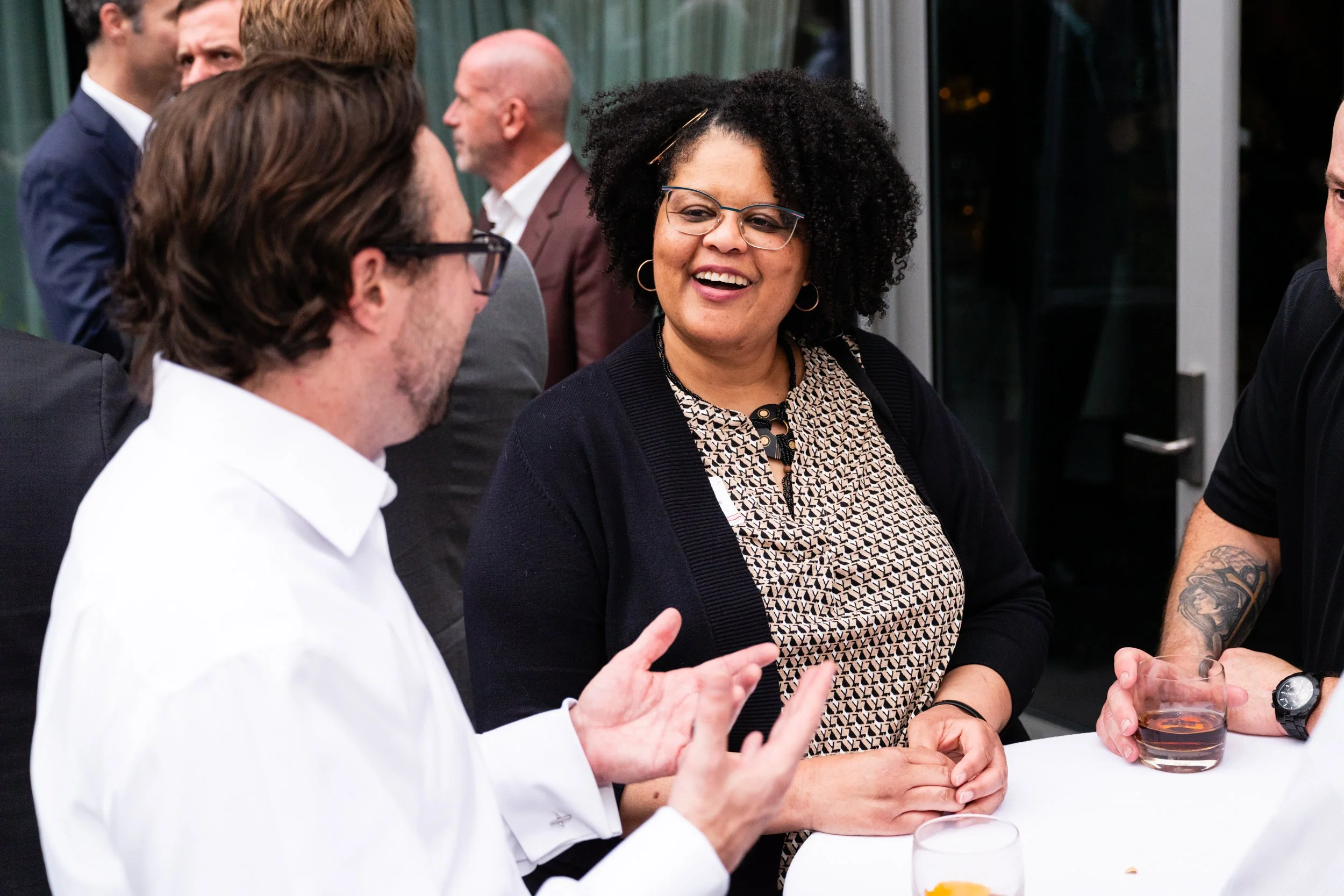 A woman with curly hair, glasses, and a patterned dress is smiling and engaging in conversation with two men at a social gathering, with other people in the background.