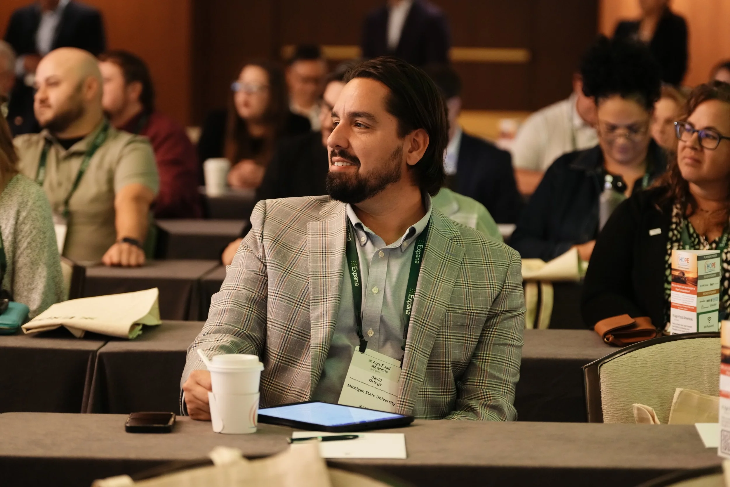A man in a plaid blazer sitting at a conference table with a coffee cup, tablet, and phone, attending a conference with other people in the background.