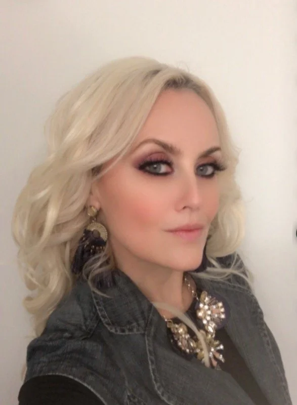Woman with blonde curly hair, makeup, earrings, jewelry, and a dark denim jacket, posing against a plain light-colored background.