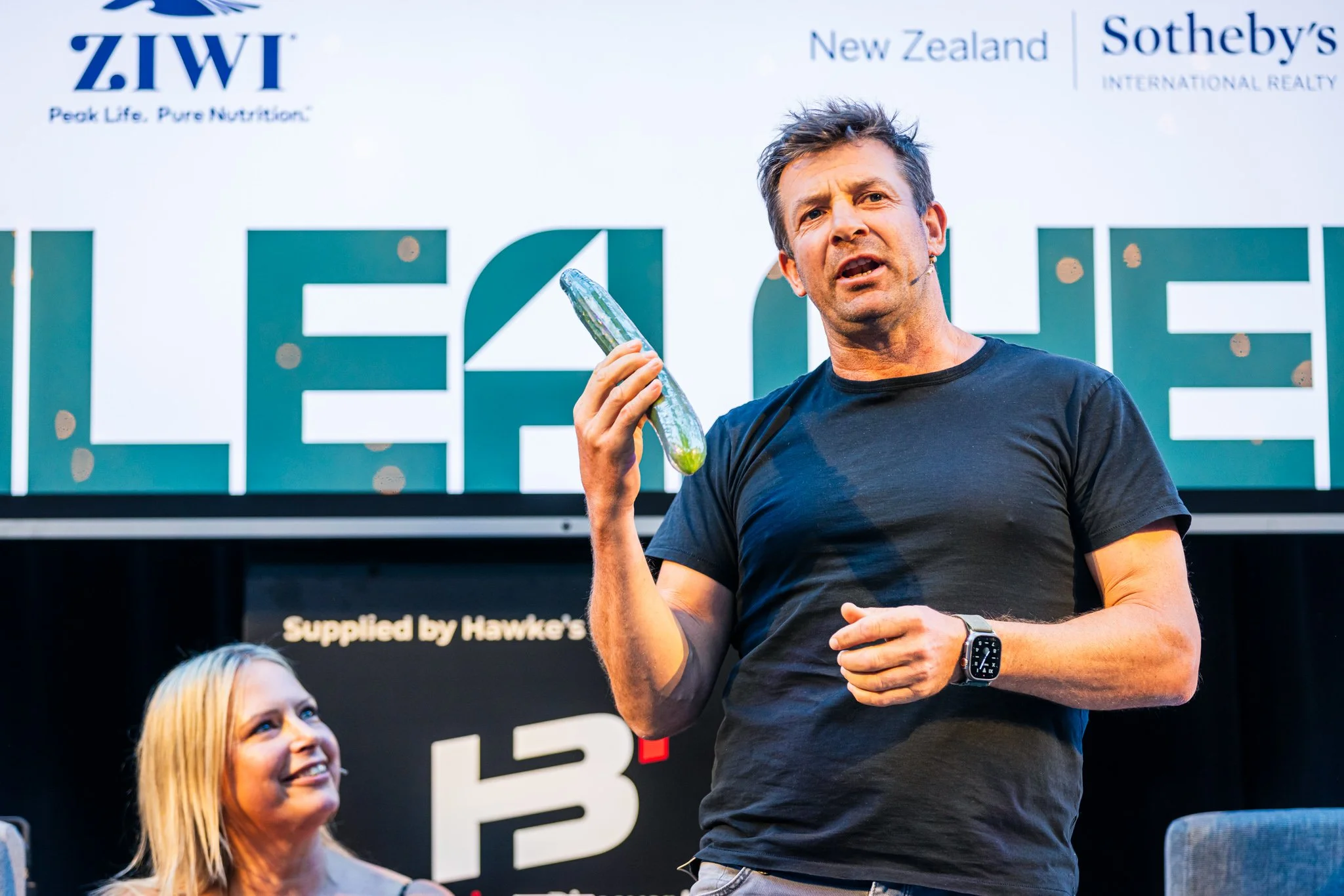 A man holding a zucchini in his right hand, speaking on stage at an event. A woman with blonde hair is sitting and looking up at him, smiling. The background has logos and text, including 'LEAP,' 'New Zealand,' and 'Sotheby's International Realty.'