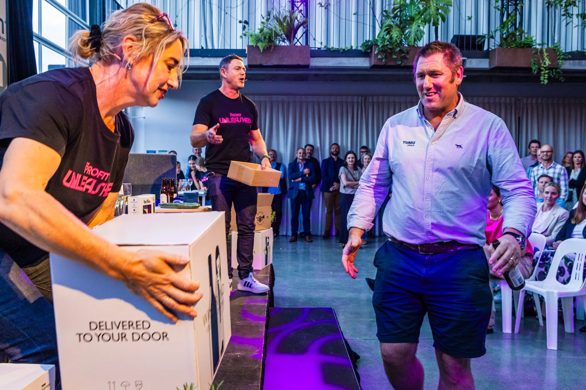 A woman and a man are at an indoor event, with the woman on the left holding a box labeled 'Delivered to Your Door' and the man on the right smiling at her. There are people watching in the background.