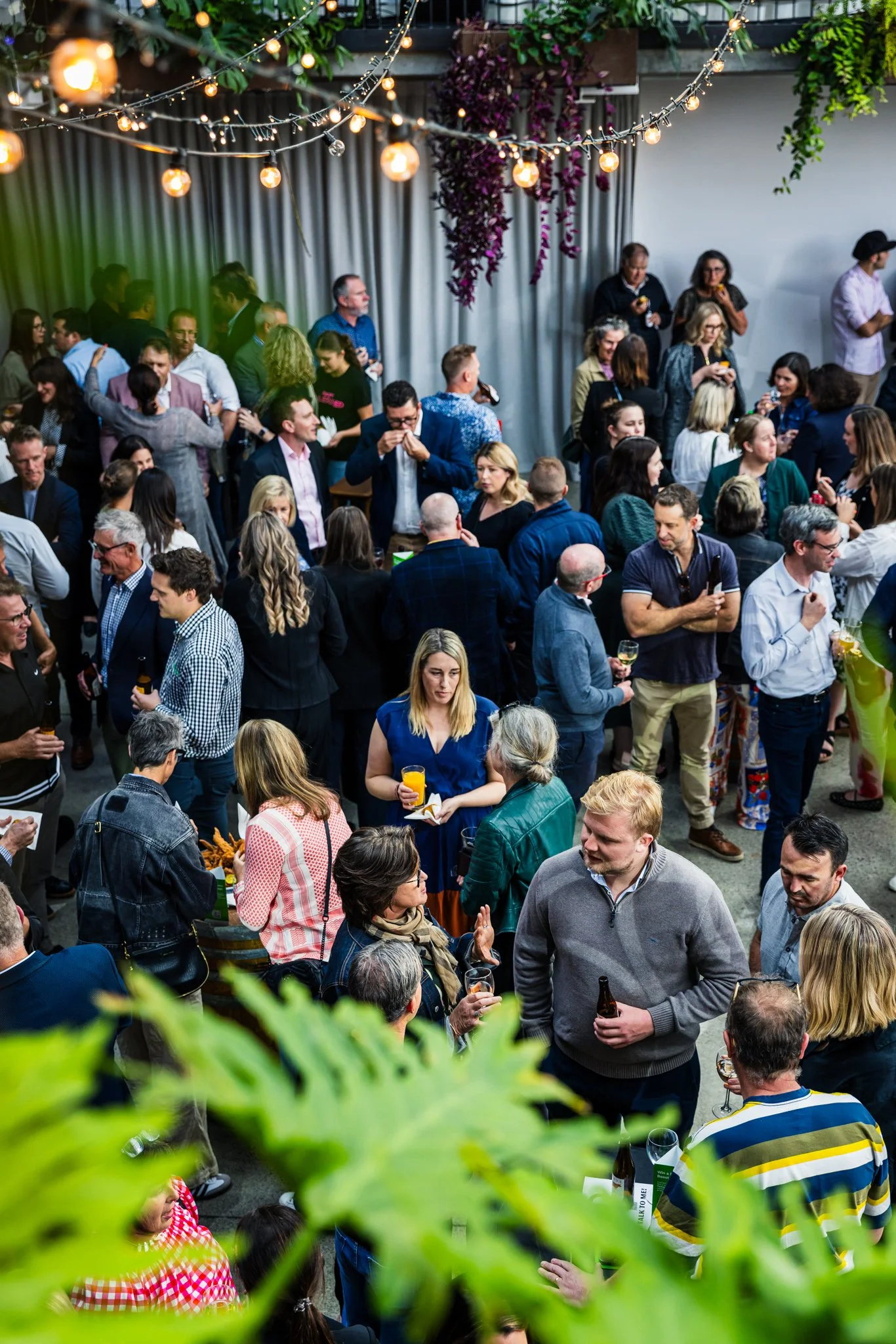 Crowd of people at an indoor party or social gathering, with string lights hanging overhead and greenery in the foreground.