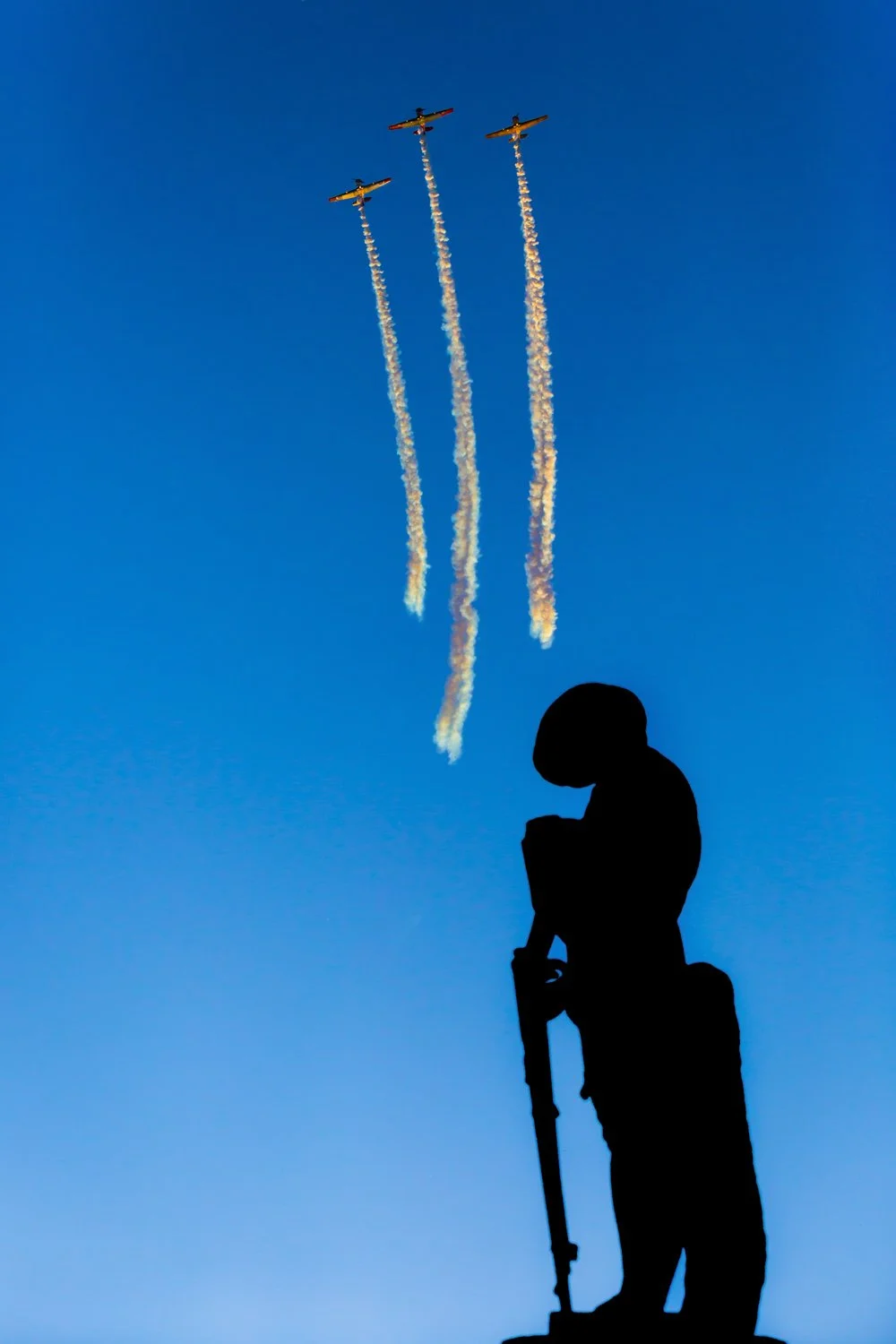 Silhouette of a person holding a rifle, watching three aircraft performing an air show against a clear blue sky with smoke trails.