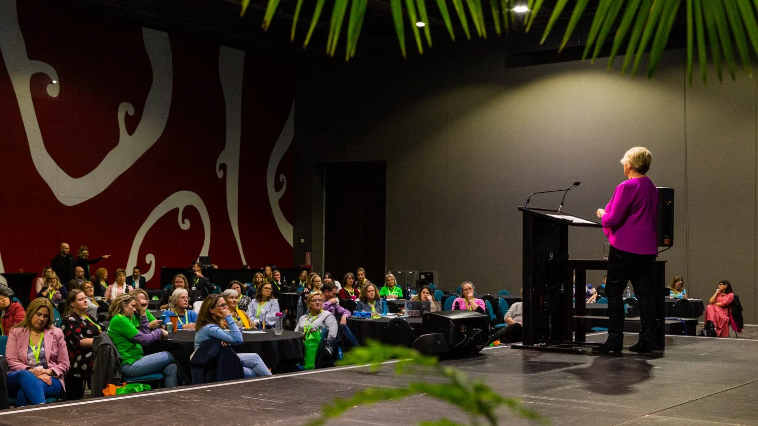 A woman in a magenta blazer giving a presentation to an audience in a conference room.