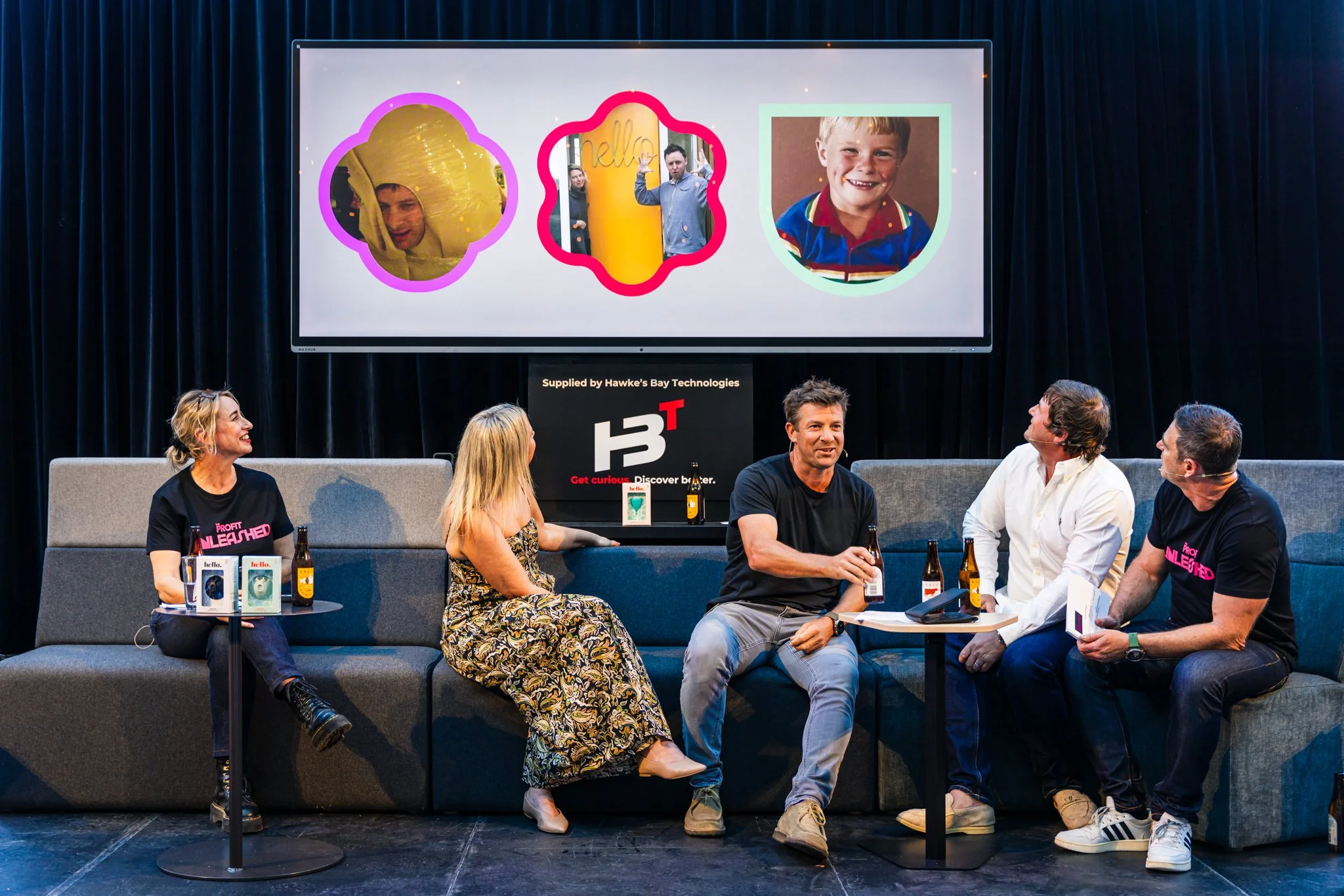 People sitting on a gray couch with drinks, having a discussion on a stage, with a large screen behind them displaying three images of children with colorful borders.
