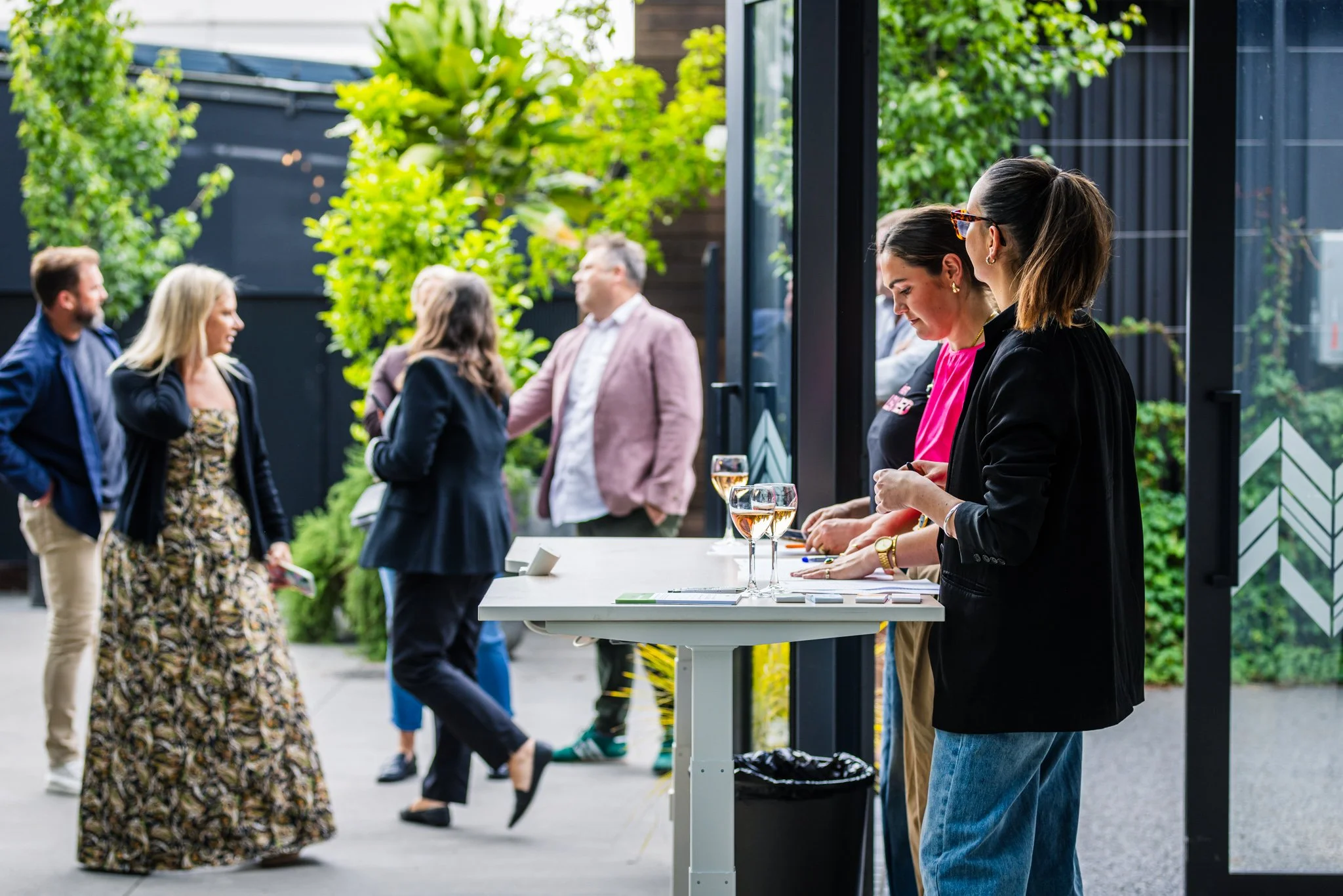 People socializing outdoors at a modern building with green foliage, some standing at a registration or information table with glasses of drinks, others engaged in conversation.
