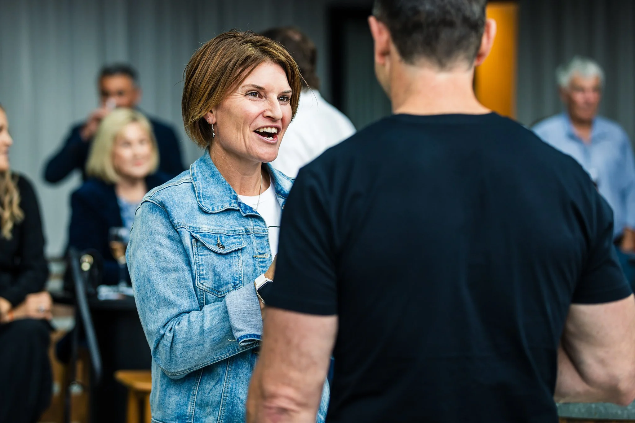 Two people engaged in conversation at a social gathering with several others in the background.