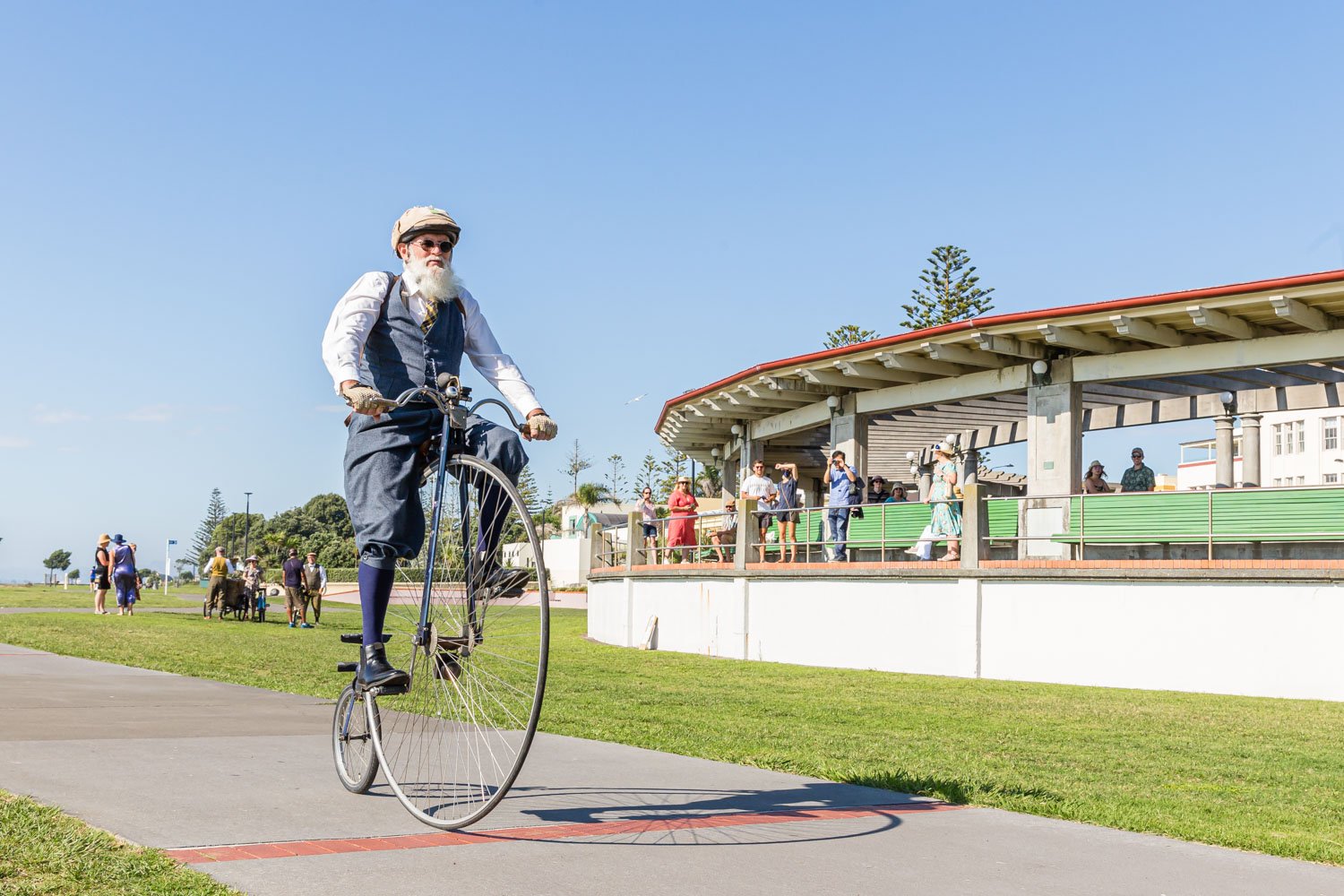 A man dressed in vintage clothing riding a tall bicycle with a small wheel at the front and a larger wheel at the back during daytime at a park. In the background, people are gathered on a pavilion and walking on the grass.